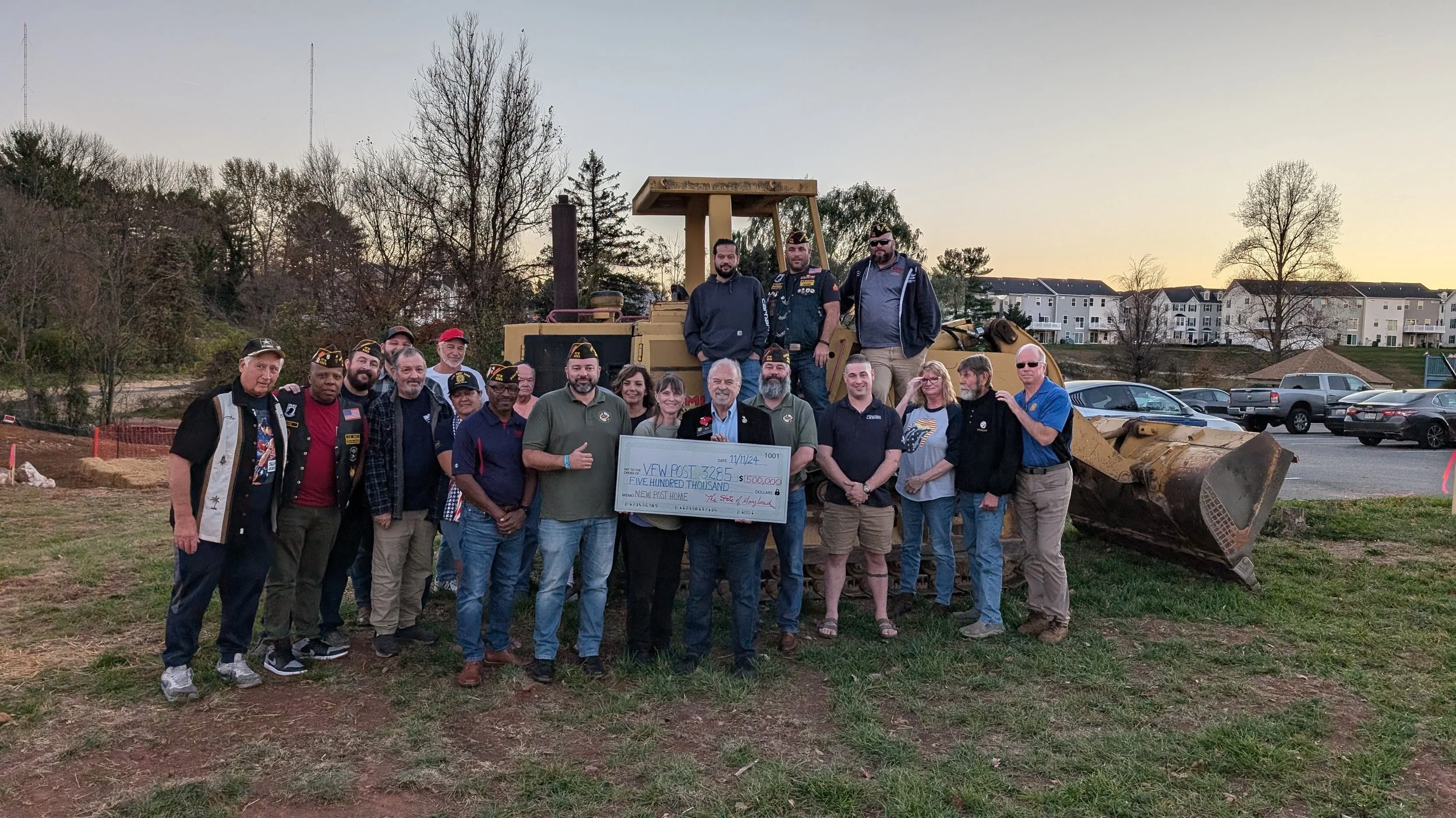 Group of people, including veterans, gathered outdoors in front of a large construction vehicle at sunset, holding a giant check for $500,000 made out to a new post home.