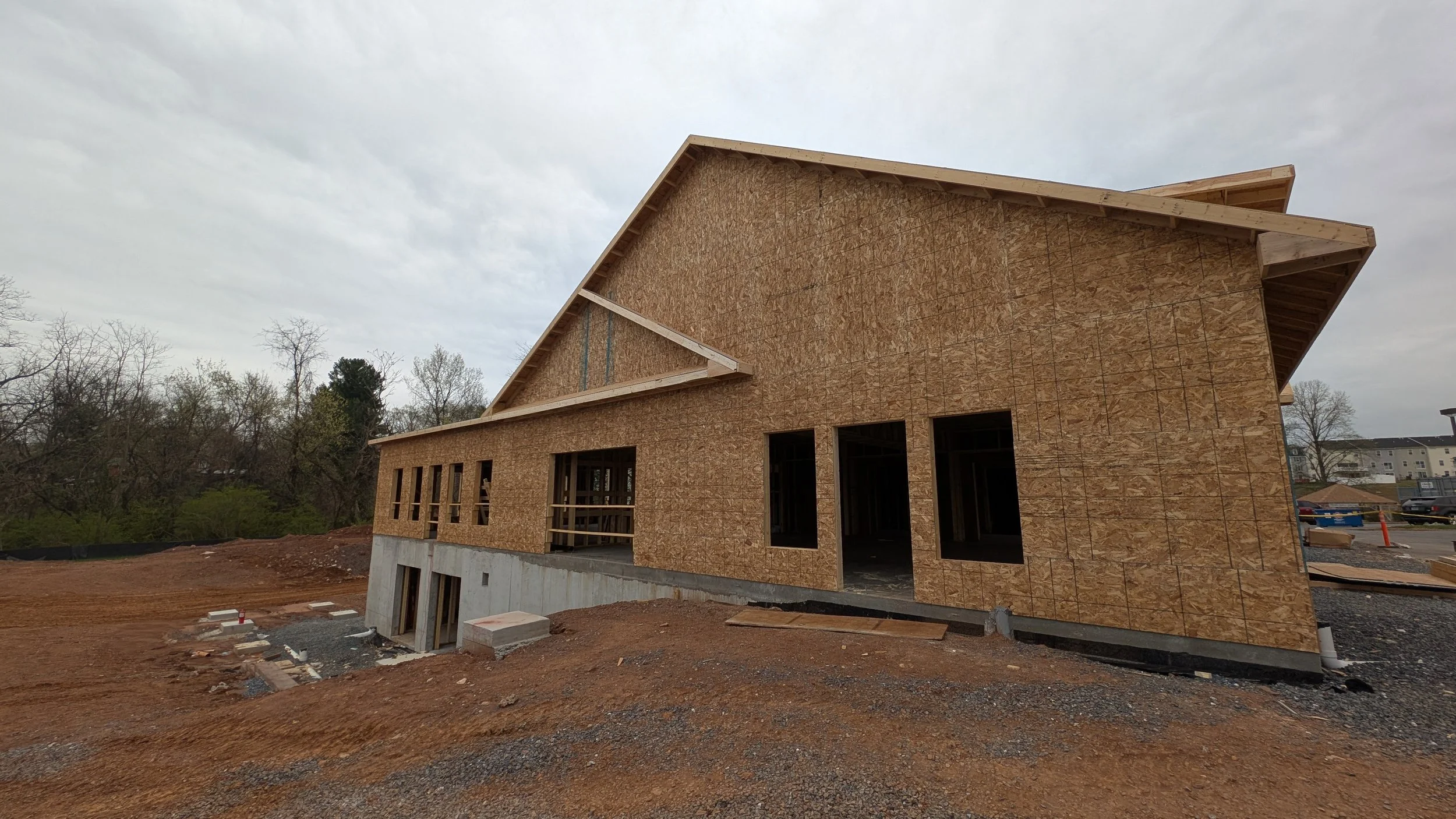 Construction site of a house with wooden framing and OSB sheathing, situated on a sloped terrain with dirt and gravel. Some windows and door openings are visible, and the house has a modern, angular roof design.