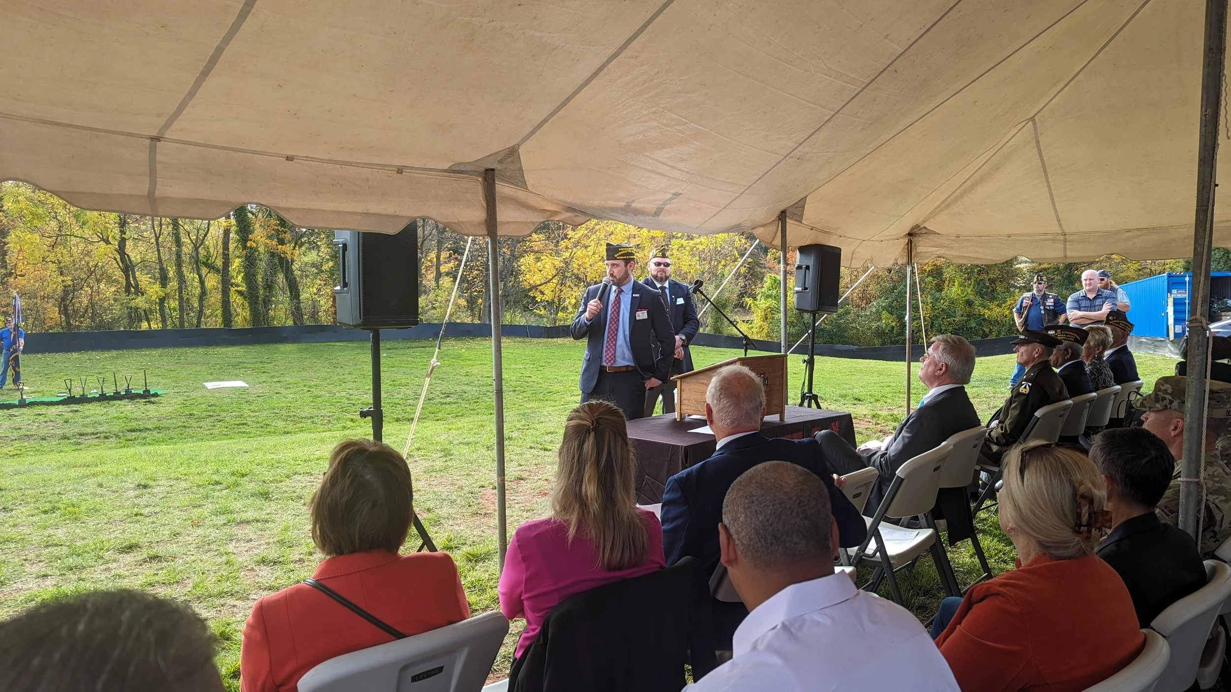 A speaker in a suit and tie speaking into a microphone at an outdoor event under a tent, with seated attendees and military personnel in uniform listening.
