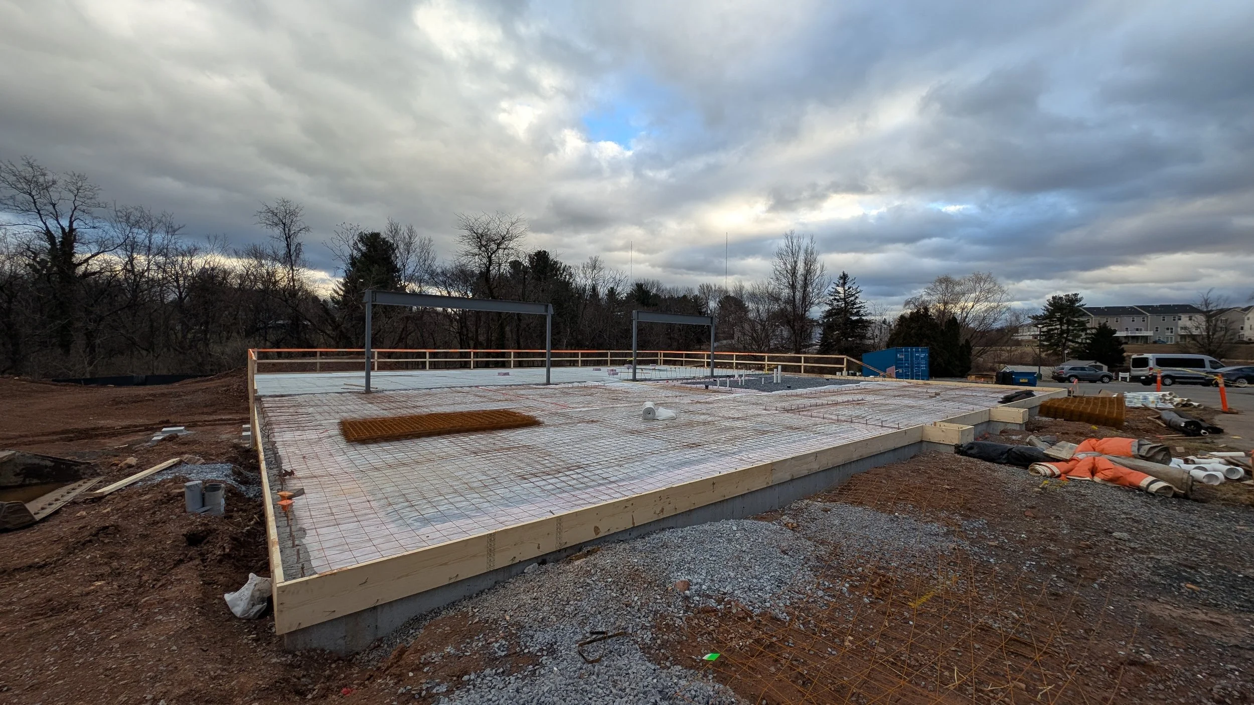 Construction site with a foundation in progress, surrounded by soil, gravel, and construction materials, under a cloudy sky.