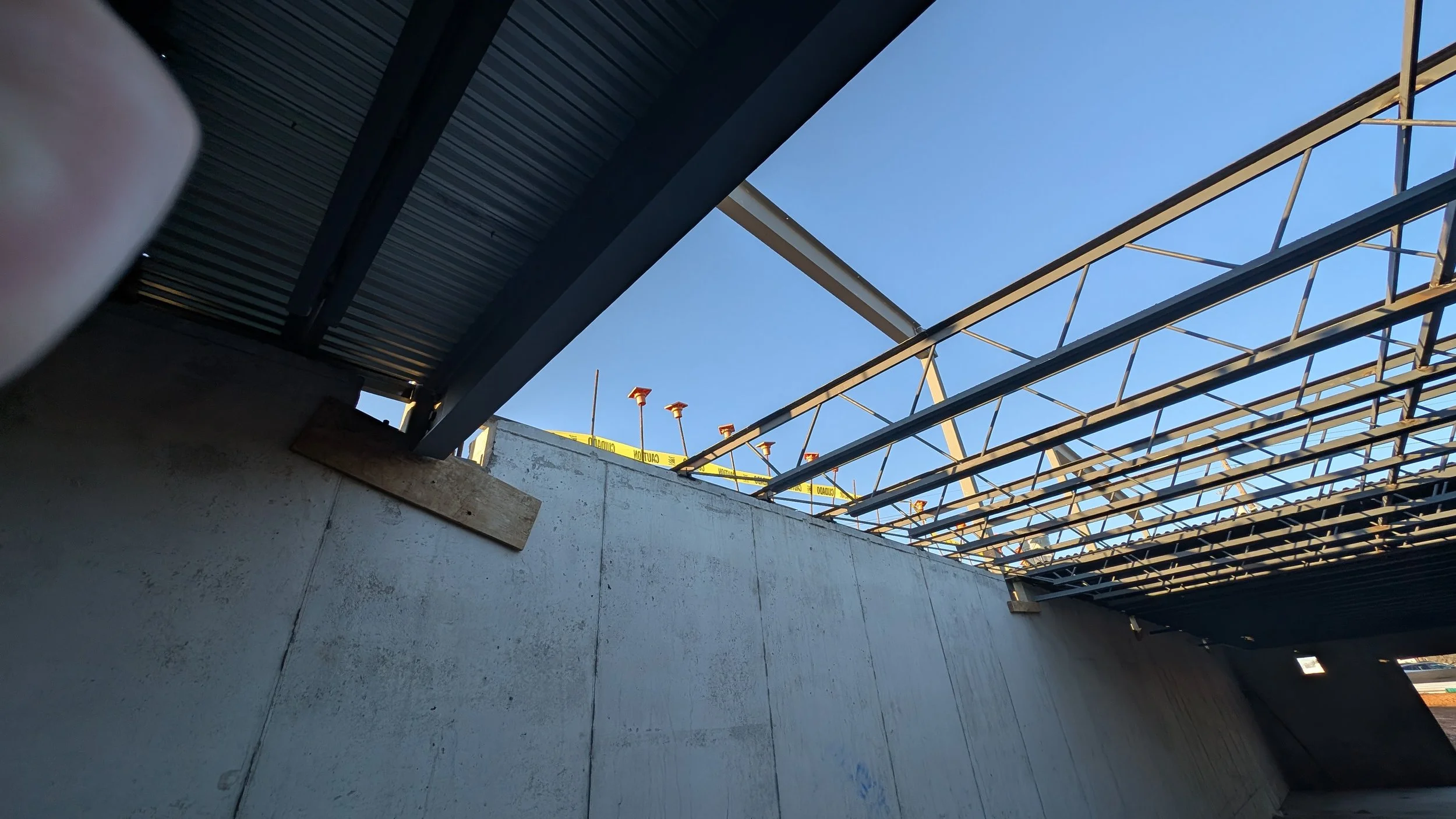Construction site with steel framework over concrete wall, yellow caution tape, and blue sky in the background.