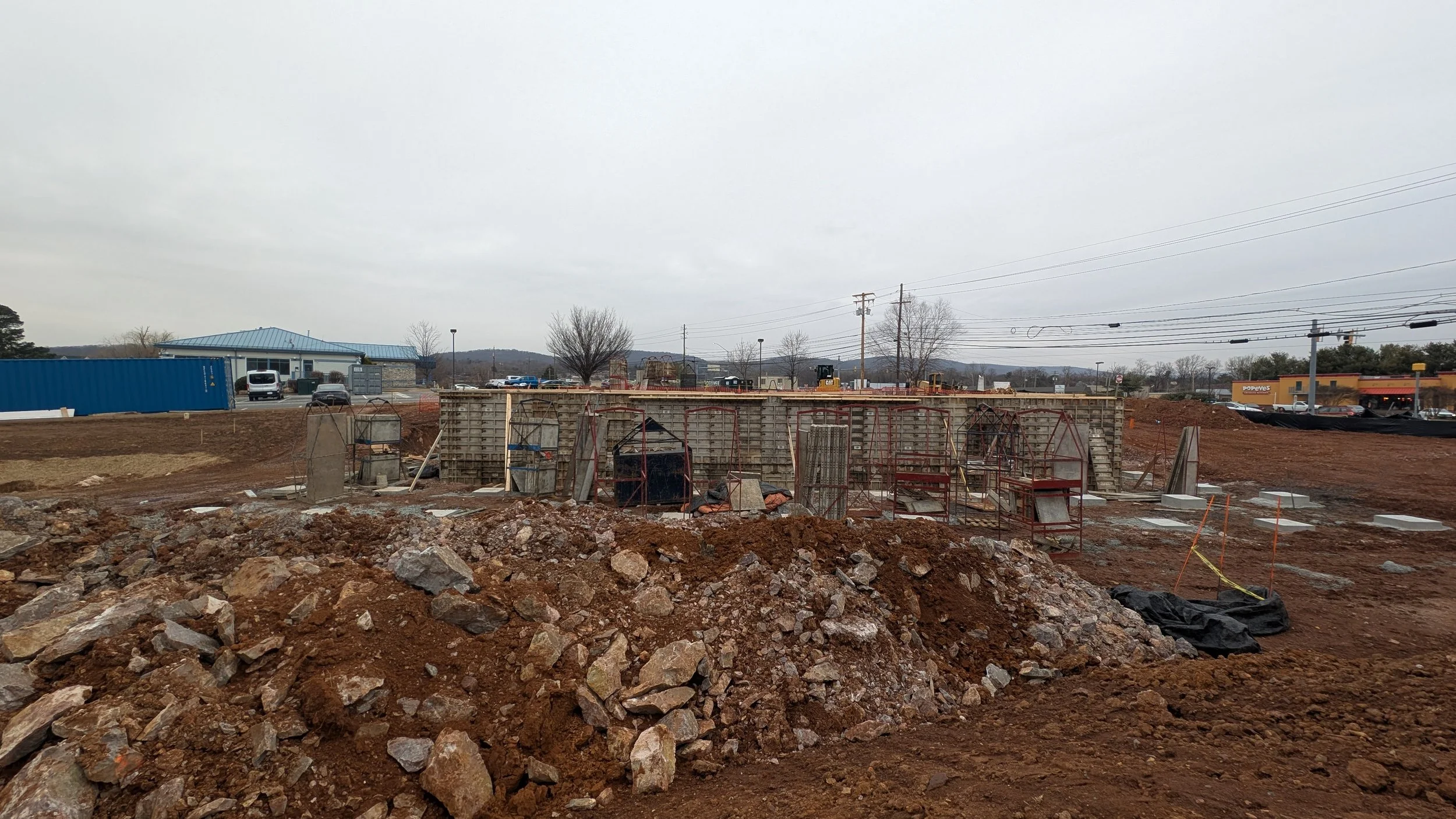 Construction site with dirt and rocks in fore, partially built concrete structure with metal reinforcements, and background showing parked cars, buildings, and power lines under cloudy sky.
