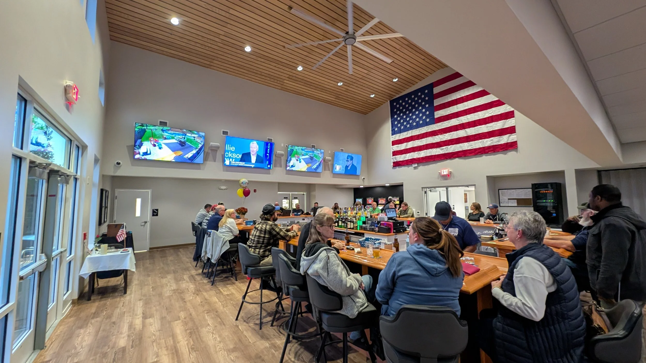 People gathered at a bar in a large, well-lit room with four television screens and a large American flag hanging on the wall.