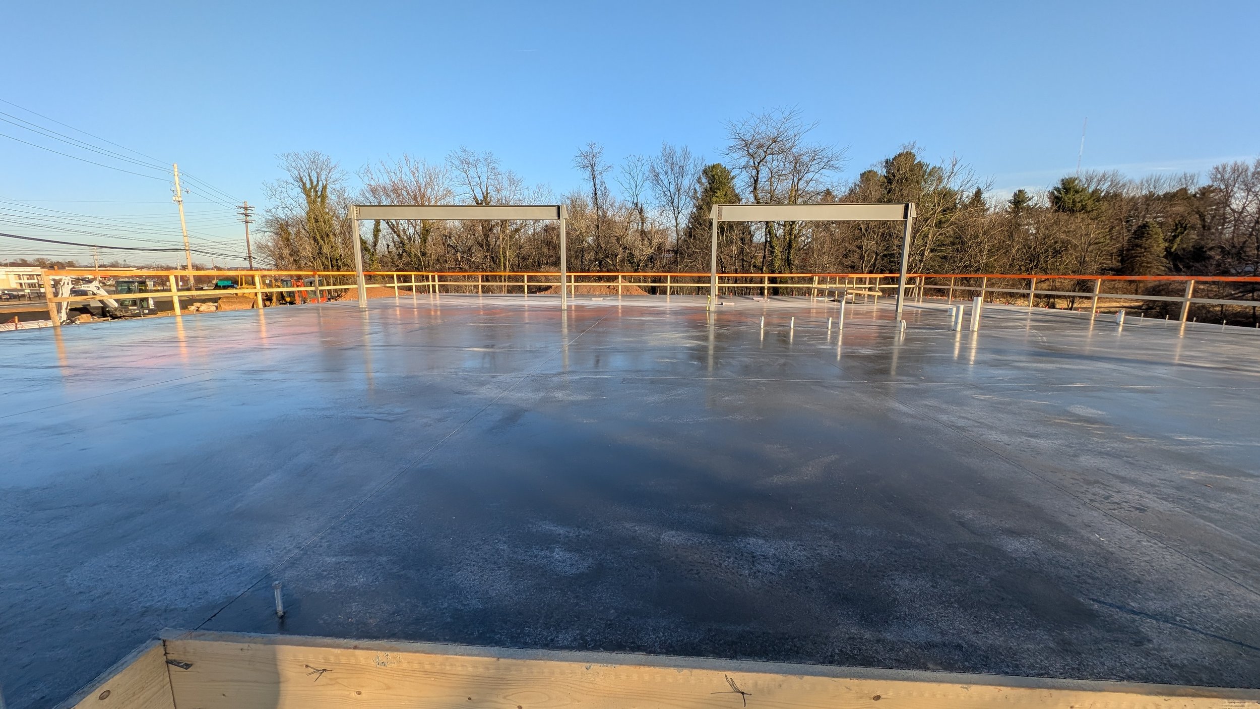 Flat rooftop with a reflective surface, surrounded by a safety railing, and metal framing structures against a backdrop of trees and a clear blue sky.