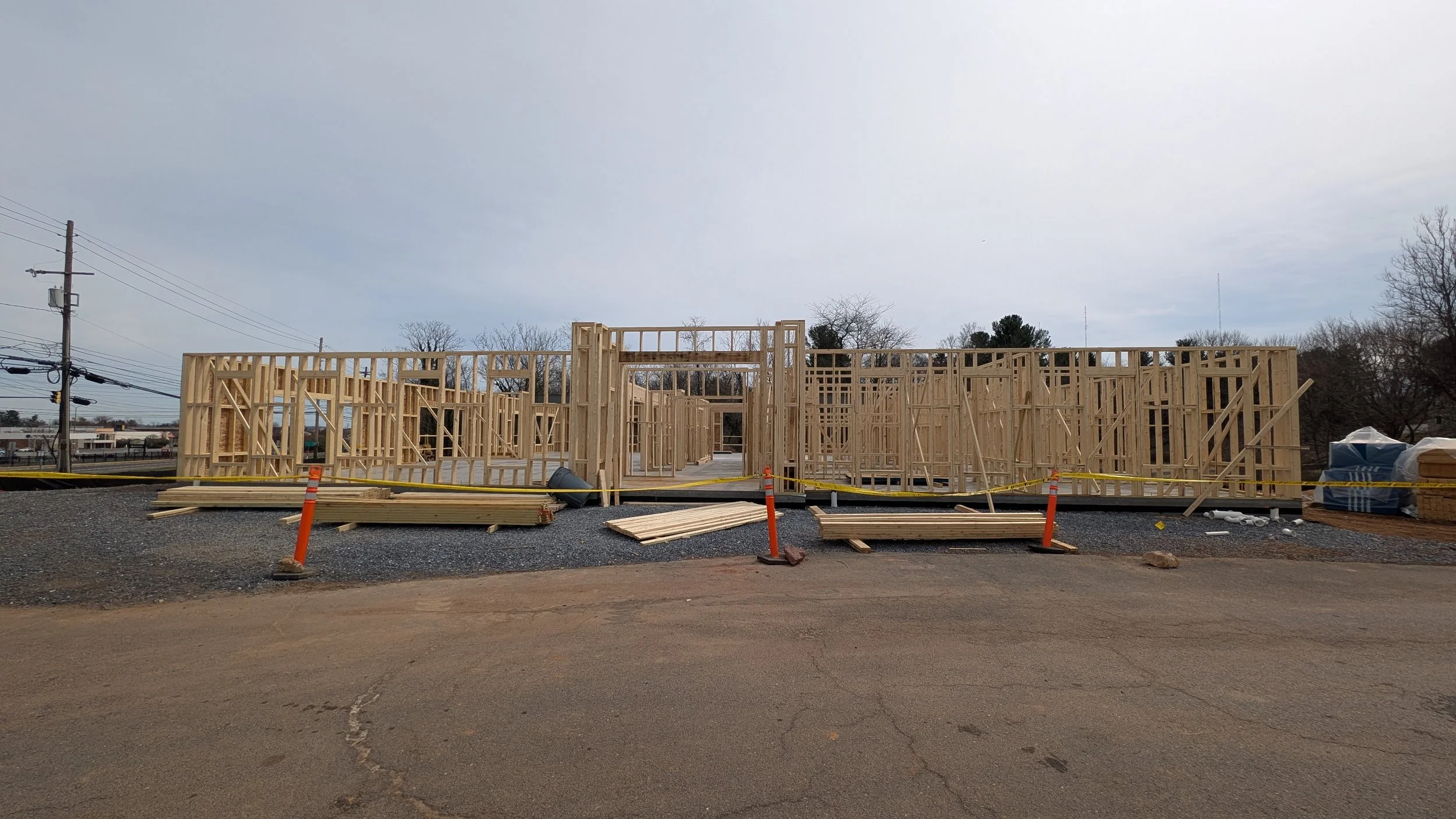 Wooden framing of a building under construction on a gravel lot, with orange construction barriers and stacks of lumber nearby, overcast sky in background.