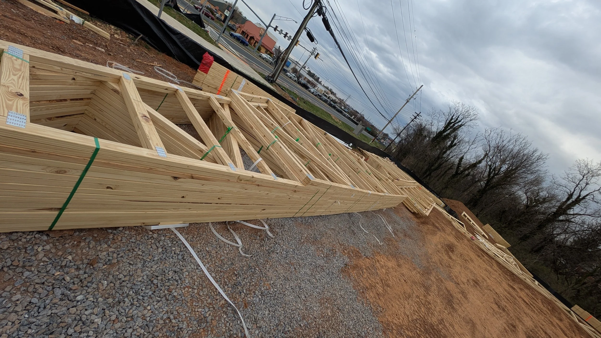 Construction site with wooden framing for a building, gravel ground, overcast sky, utility poles, and trees in the background.