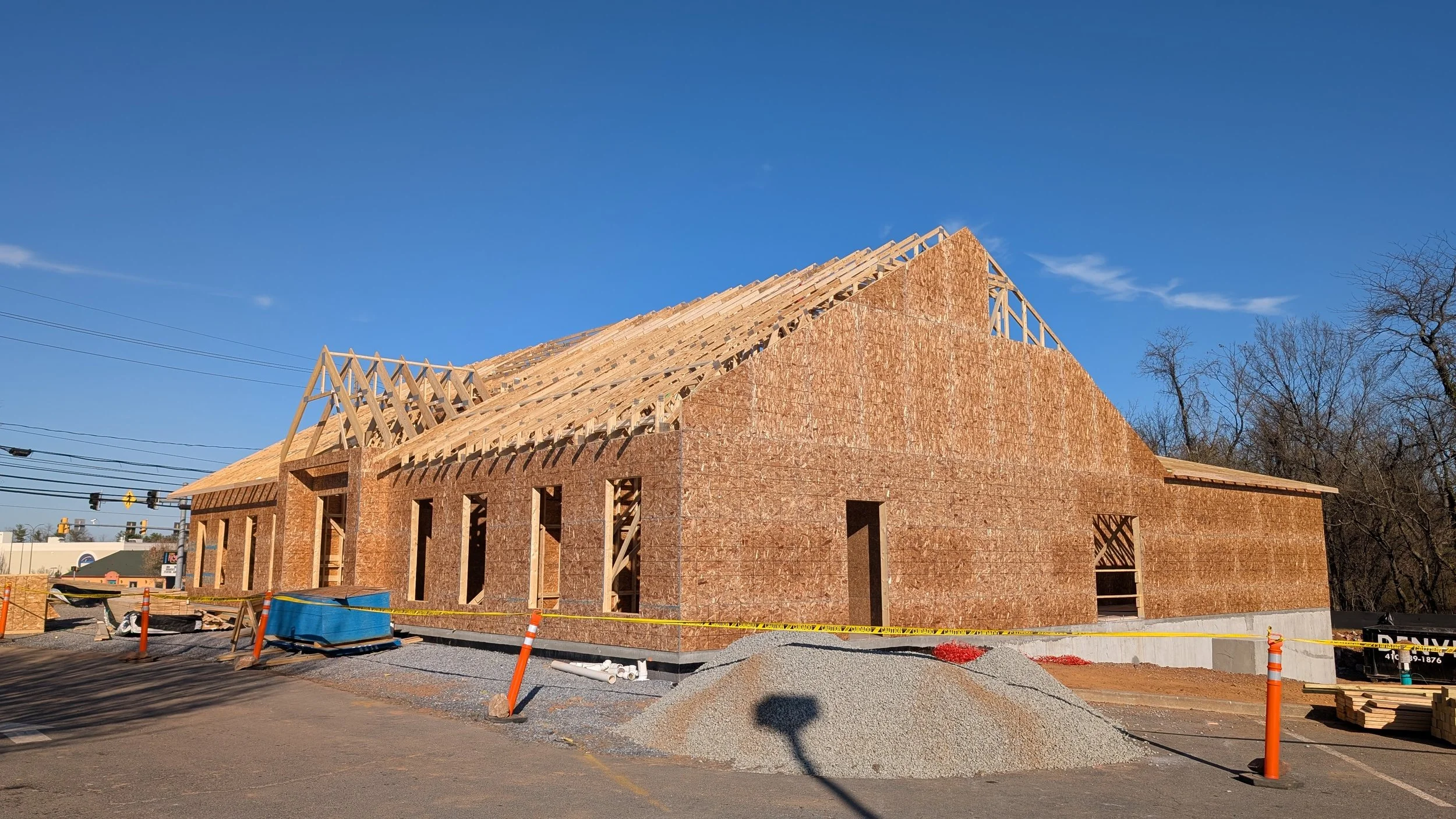 A house under construction with a wooden framework and partially built brick walls, situated on a construction site with gravel, orange safety cones, and yellow caution tape, against a background of leafless trees and a blue sky.