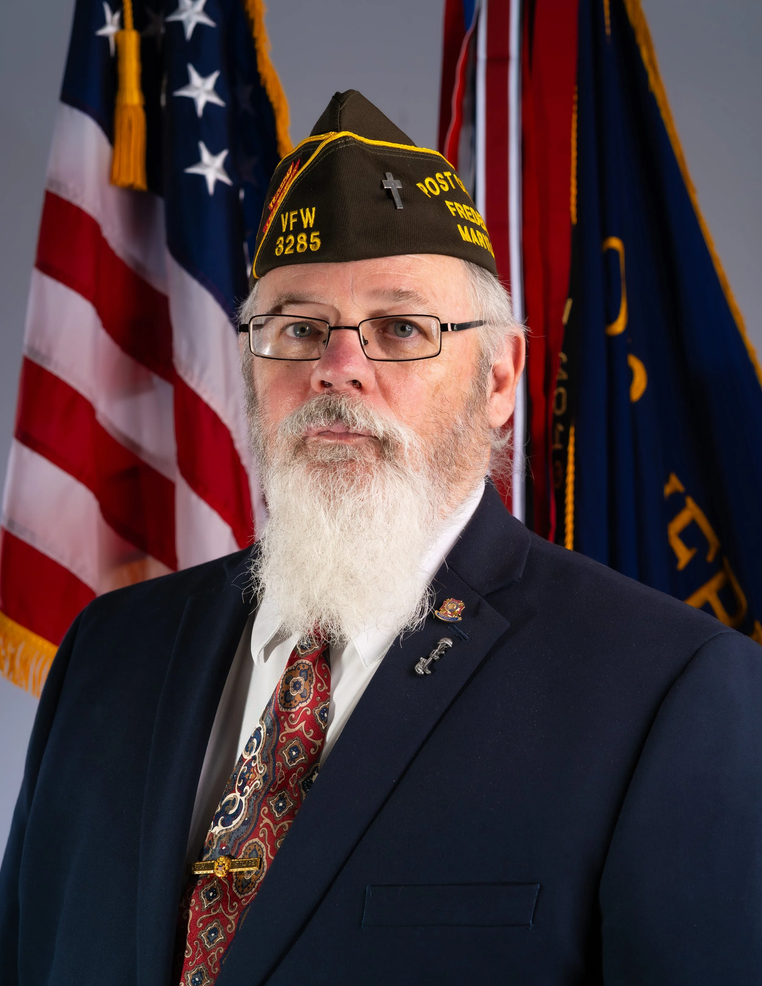A man wearing glasses and a navy blue shirt with a fire department insignia. He has a light goatee and is wearing a black cap with yellow embroidery and a red patch. Behind him are parts of an American flag and a yellow wall.