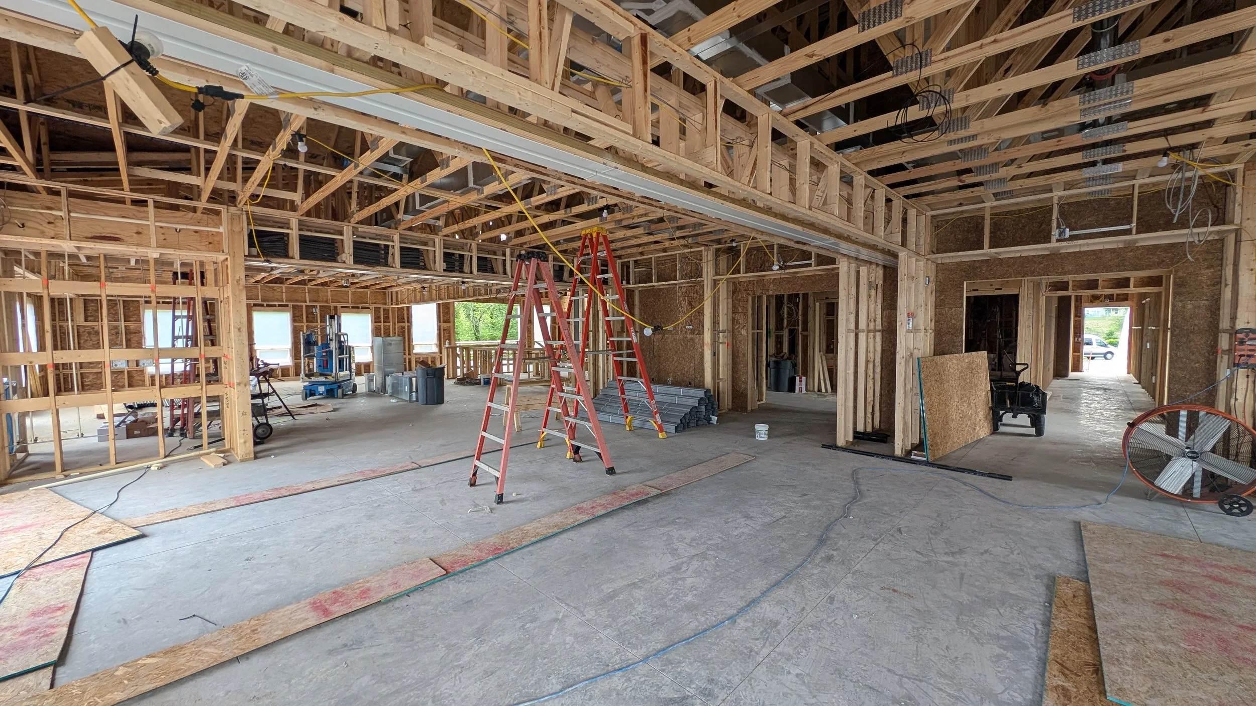 Interior of a building under construction with exposed wooden framing, construction tools, ladders, and building materials scattered around.