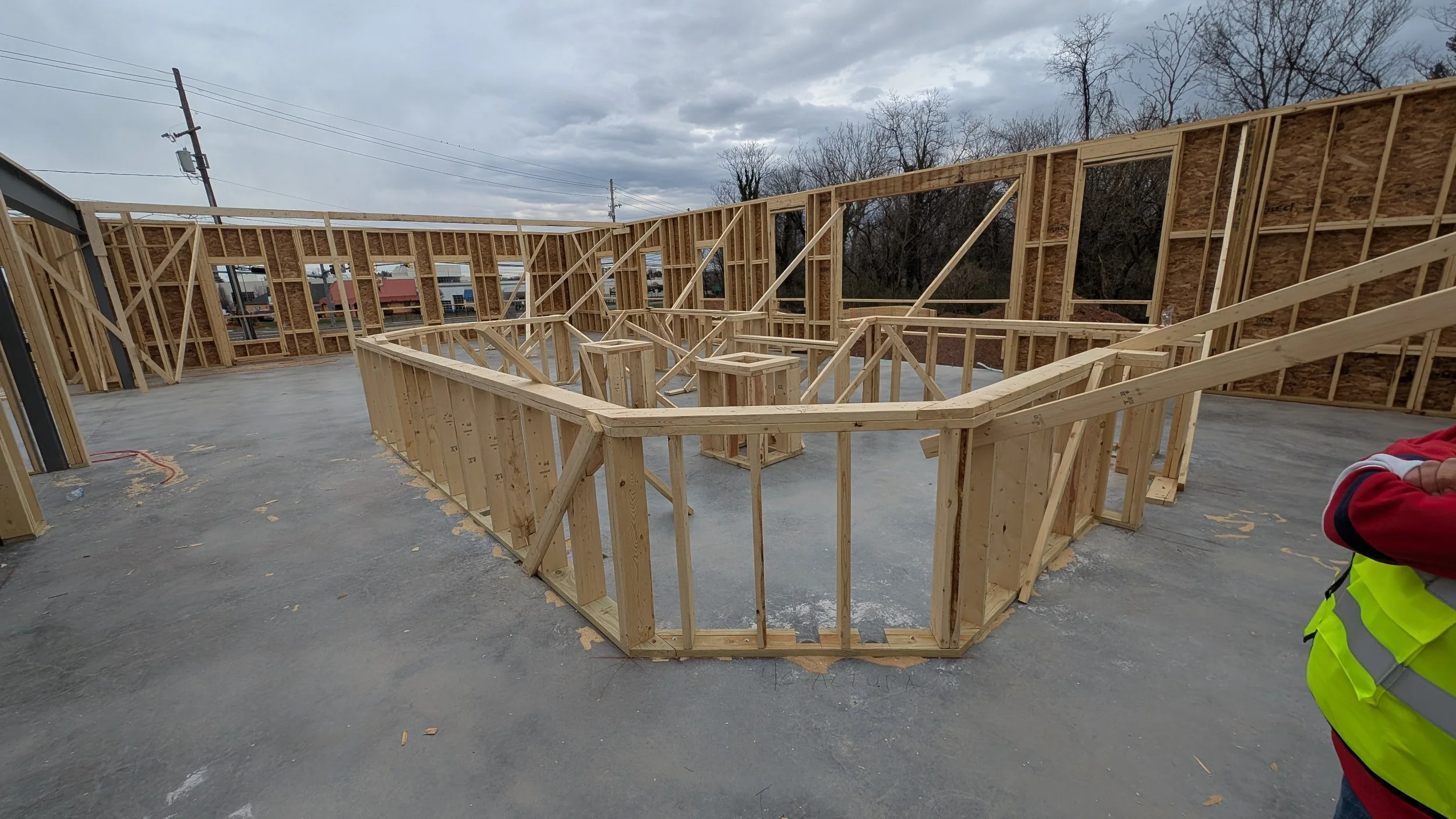 Wooden framing of a building under construction on a concrete foundation with an overcast sky. A person in a yellow safety vest and red jacket is partially visible on the right.