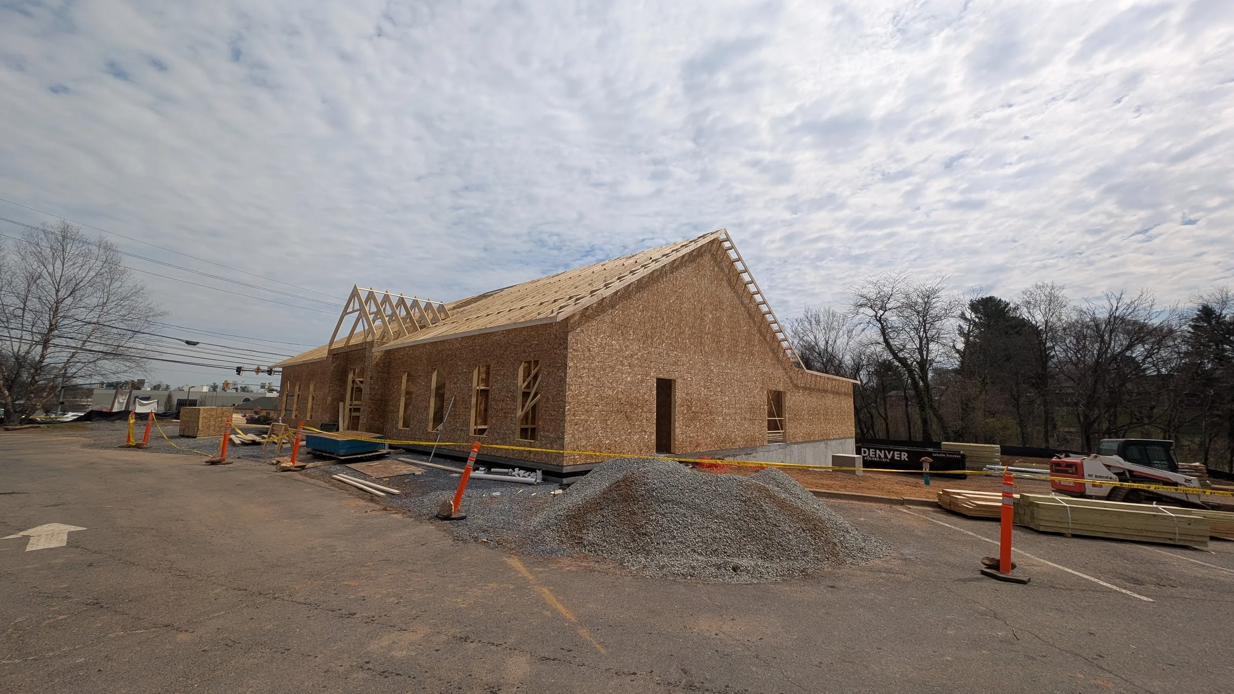 A house under construction with wooden framing on the roof, brick walls, construction materials, and orange safety cones in a parking lot area, with trees and a partly cloudy sky in the background.