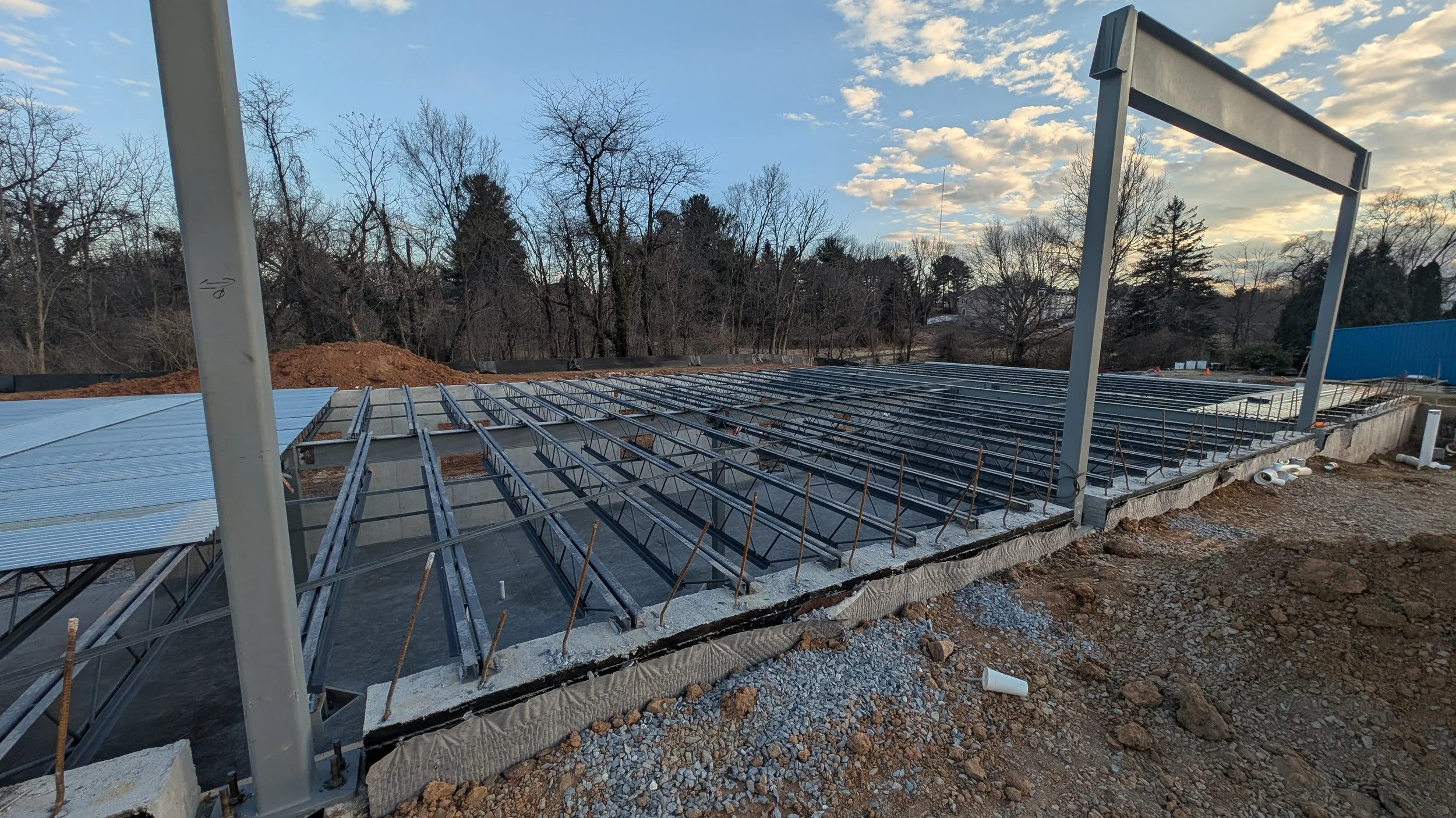 Construction site with metal framework for a building's foundation, surrounded by dirt and gravel, with trees and a cloudy sky in the background.