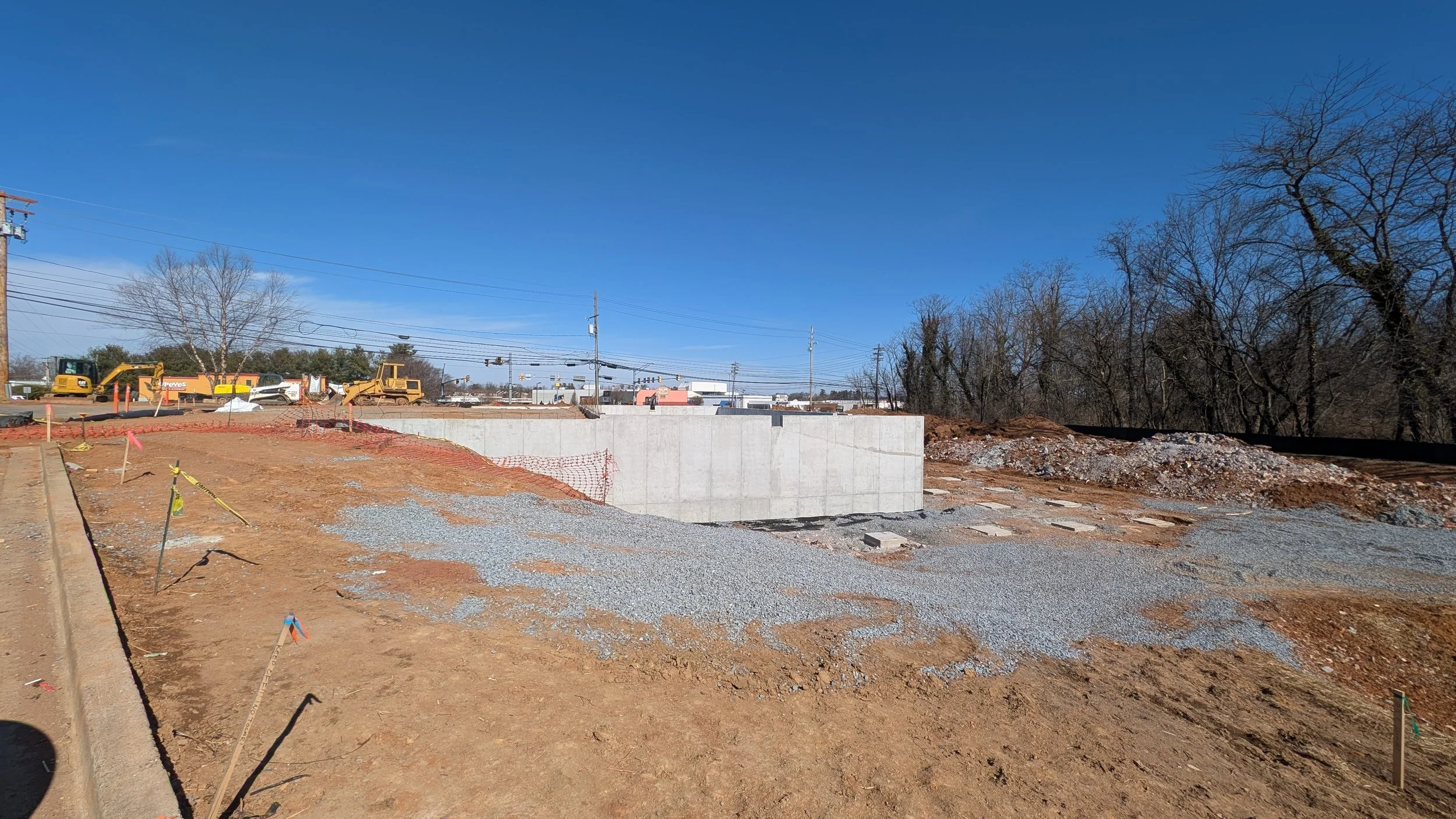 Construction site with a concrete wall, gravel, construction machinery, and trees in the background under a clear blue sky.