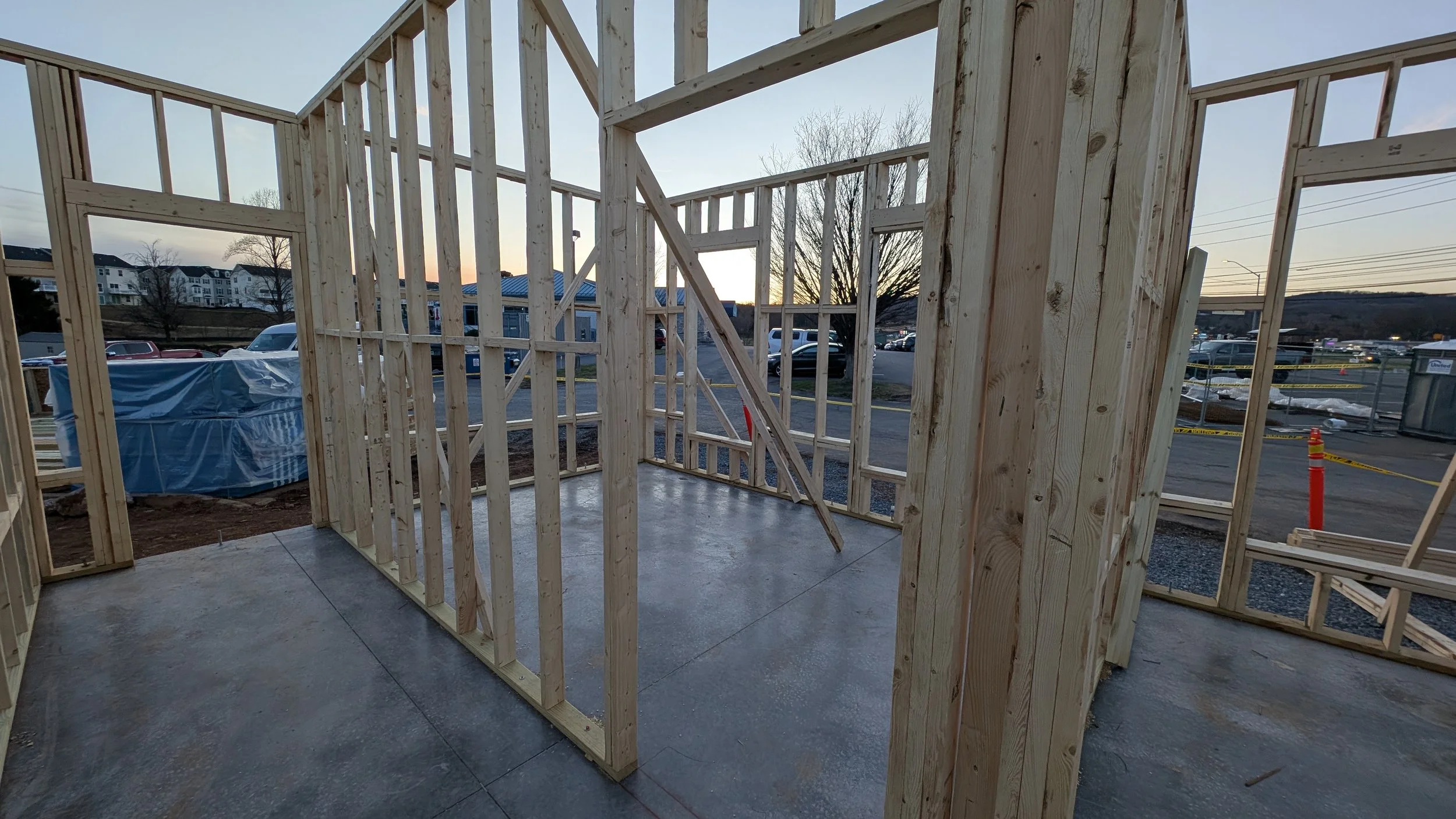 Wood framing for a building under construction at sunset, with parking lot and trees in background.