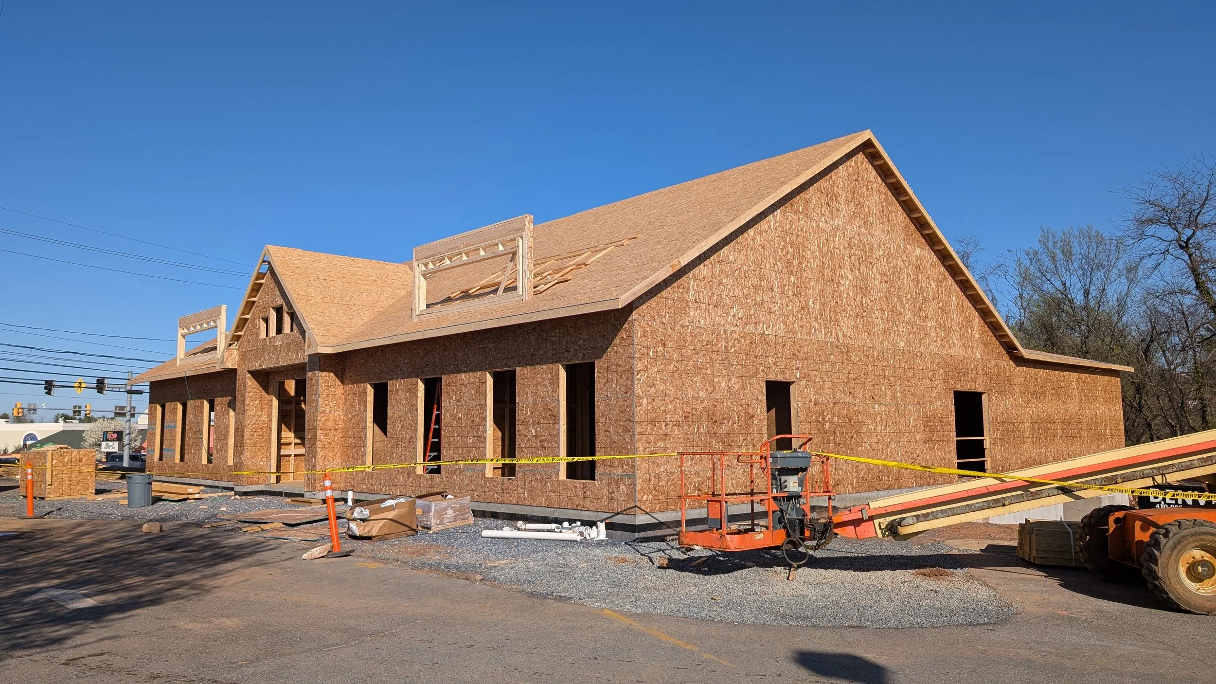 A house under construction with wooden framing and plywood sheathing, construction equipment and caution tape in the foreground, and a clear blue sky above.