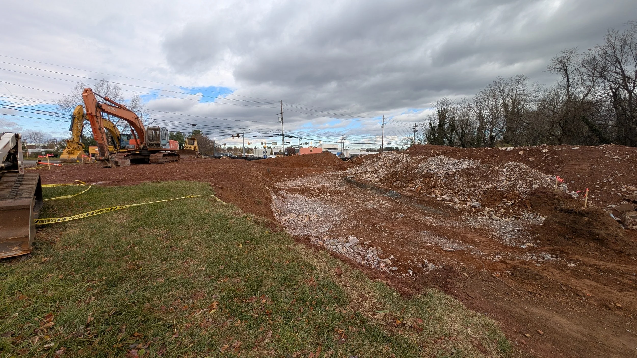Construction site with excavators on dirt and gravel, yellow caution tape, grassy area, cloudy sky, and power lines in the background.