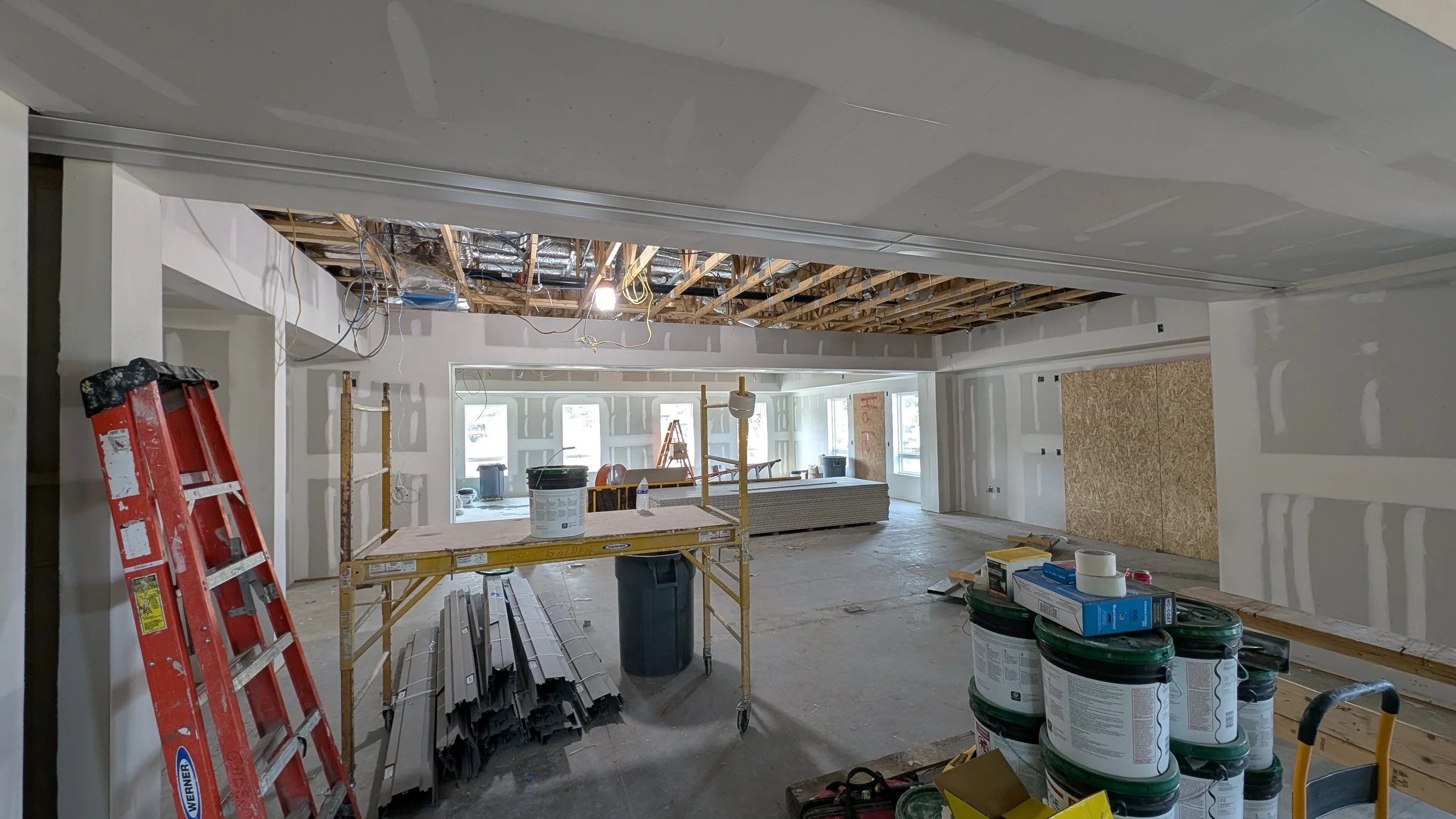Interior of a building under construction with drywall installed on walls, an exposed ceiling with wiring, construction materials, and tools scattered around.