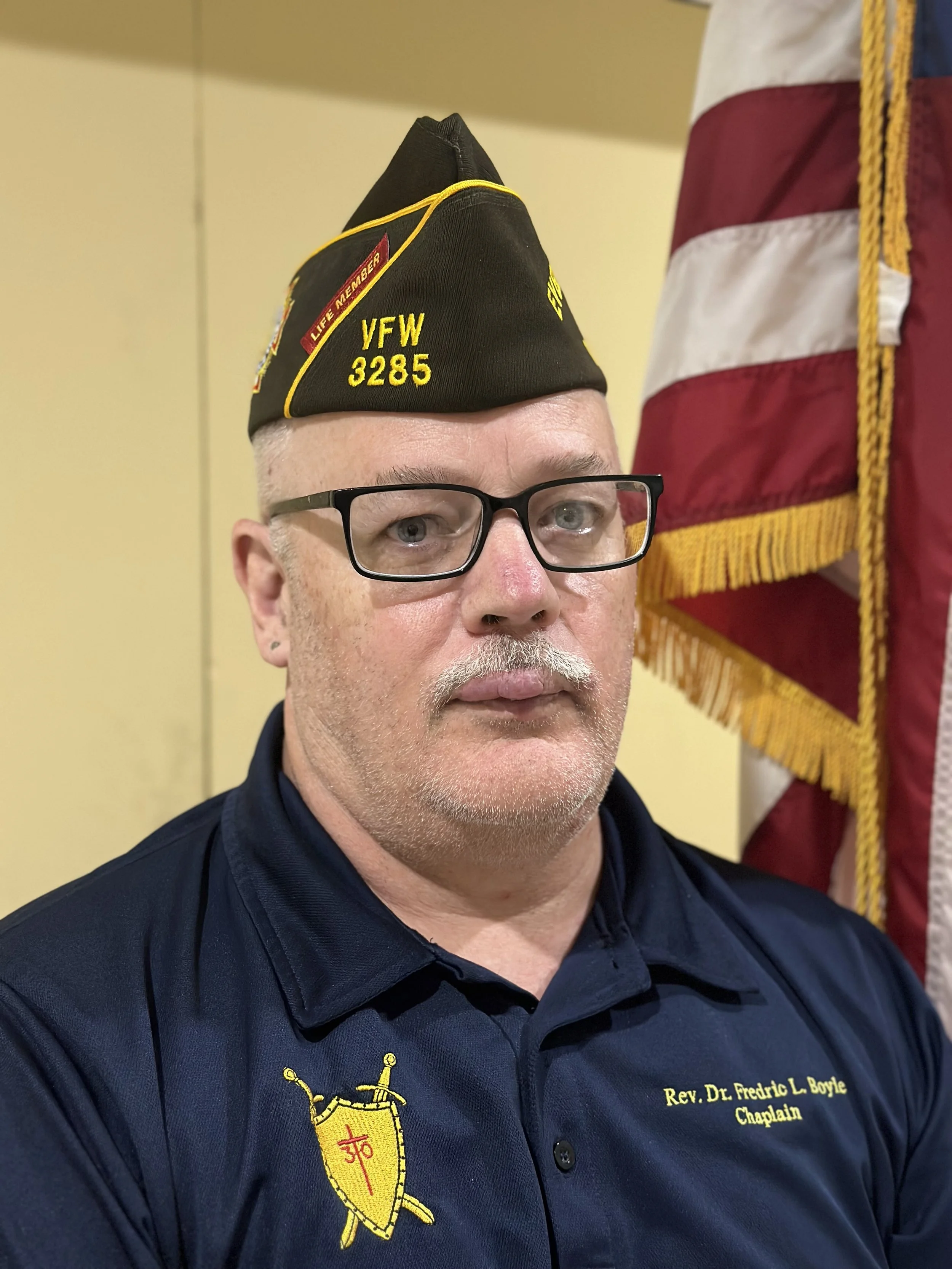 A man wearing glasses and a navy blue shirt with a fire department insignia. He has a light goatee and is wearing a black cap with yellow embroidery and a red patch. Behind him are parts of an American flag and a yellow wall.