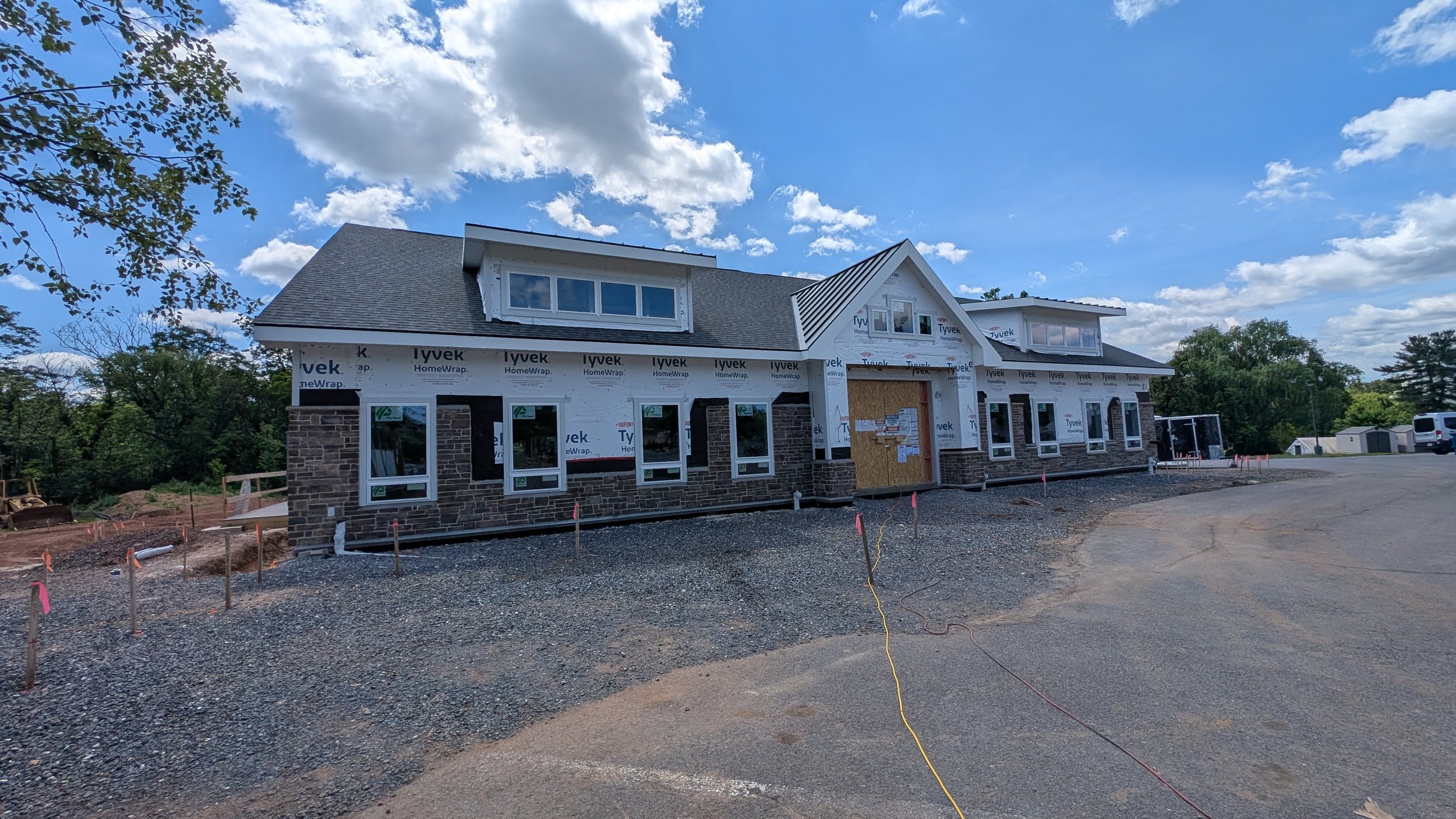 Under construction house with brick exterior and white window frames on a gravel lot under a partly cloudy sky.