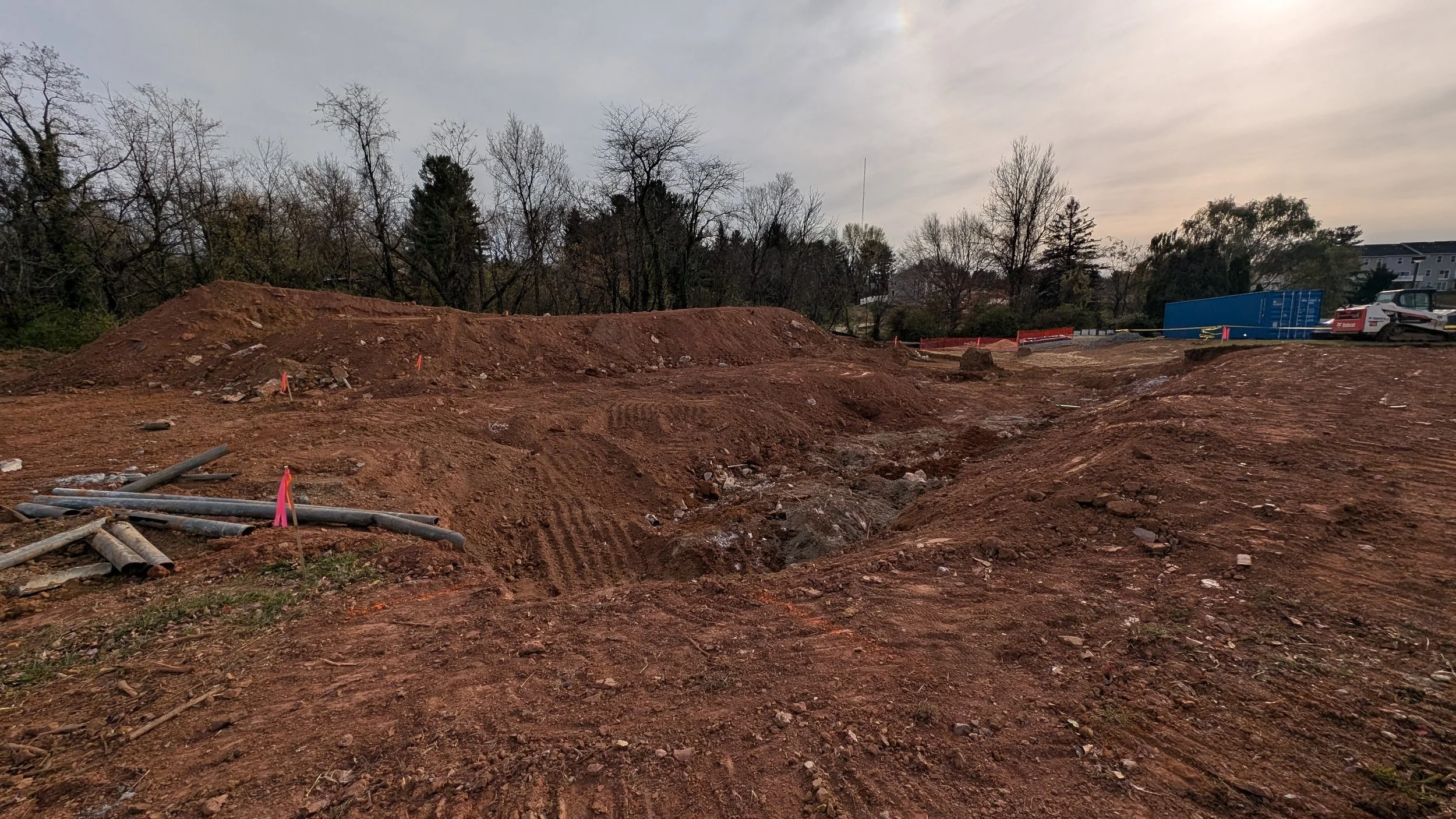 Construction site with excavated dirt and exposed pipes, surrounded by trees and buildings in the background.