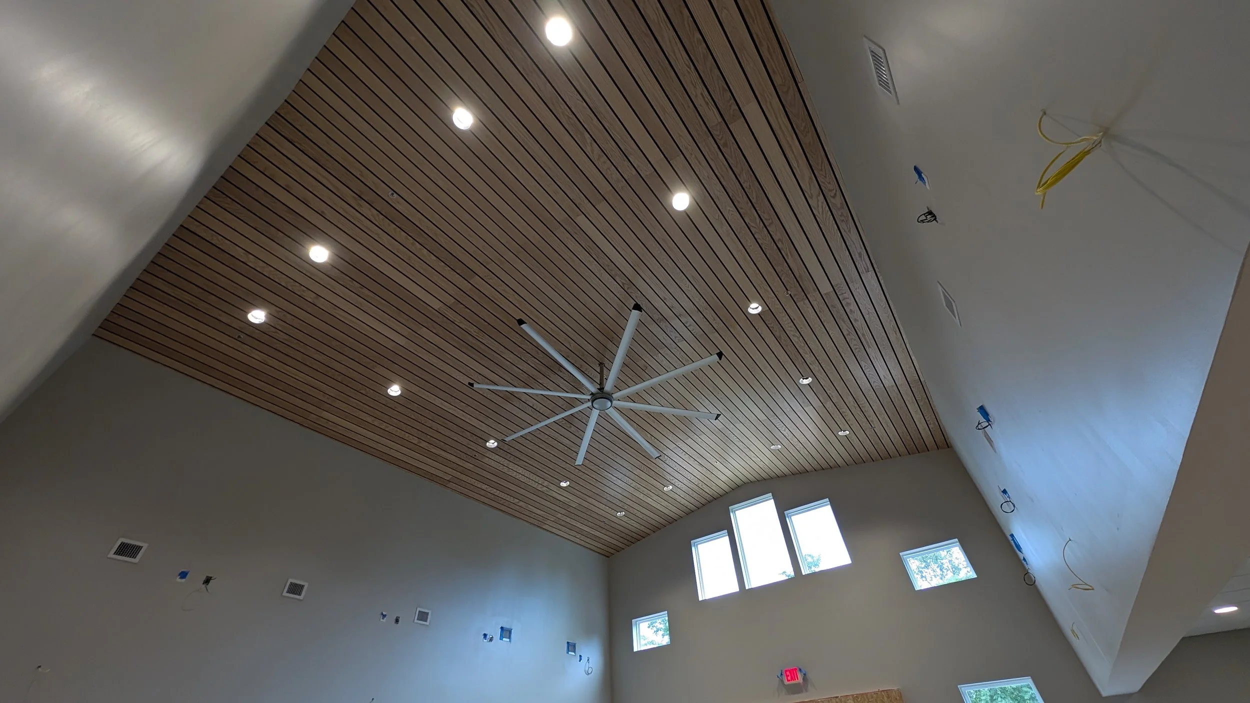 A room under construction with a wooden ceiling, recessed lighting, windows on the wall, and a large modern ceiling fan.