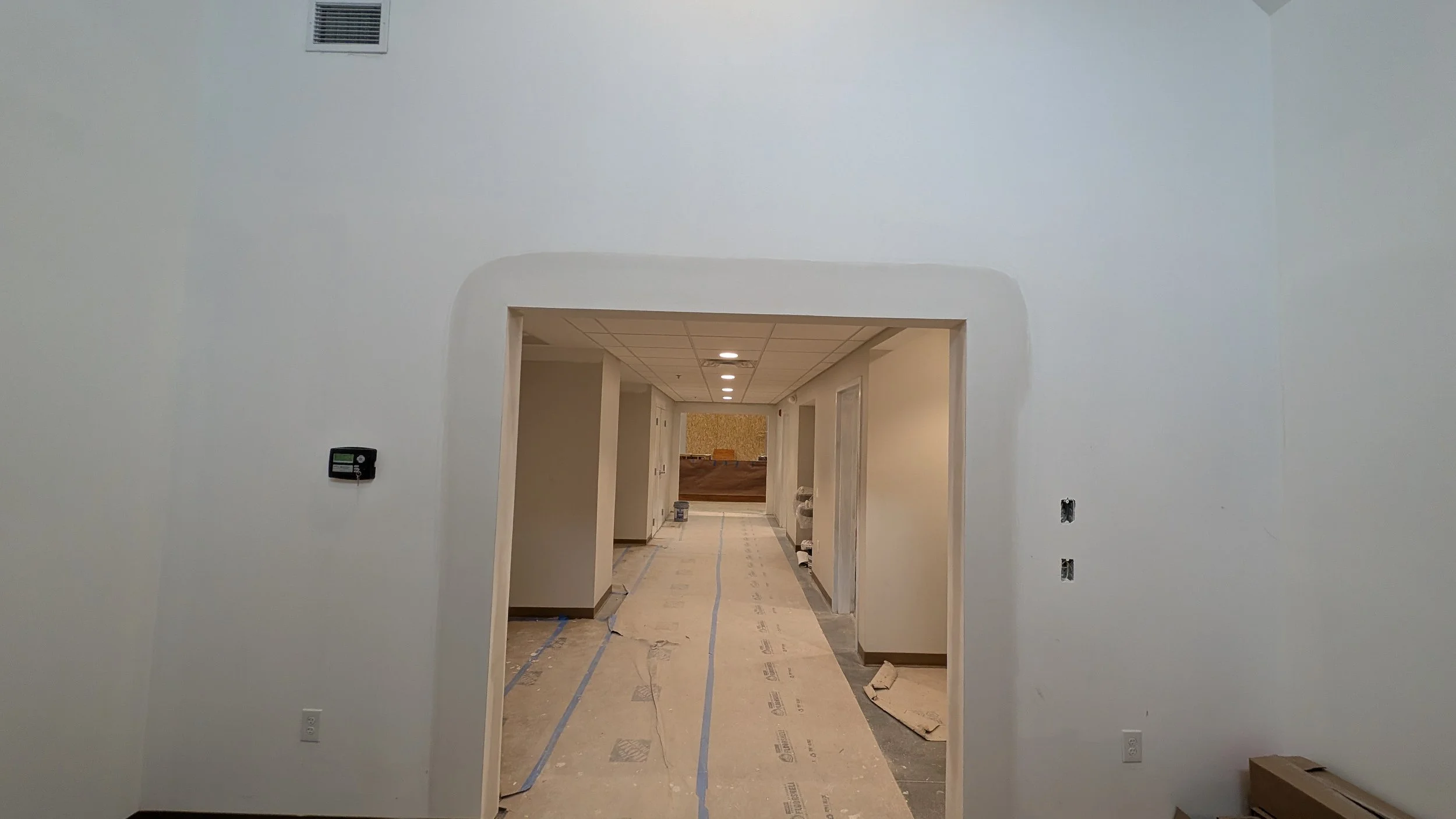 Interior hallway under construction with white walls, a partially visible doorway, and a ceiling with recessed lighting. Floor covered with protective paper and construction materials.