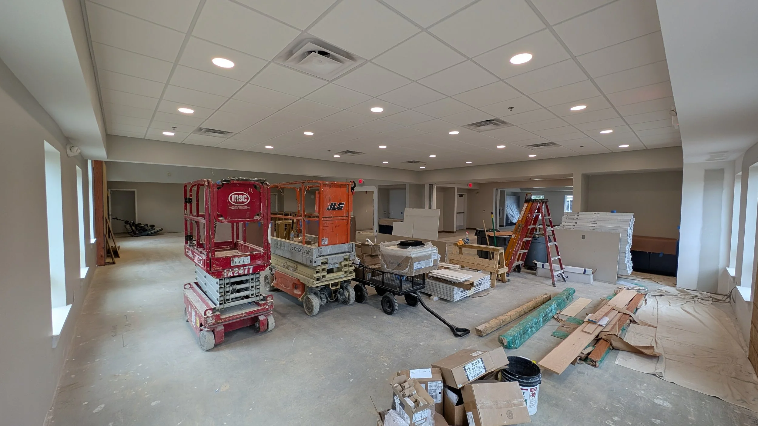 Interior of a building under construction with construction equipment, tools, and materials scattered across the floor, and new lighting installed on the ceiling.