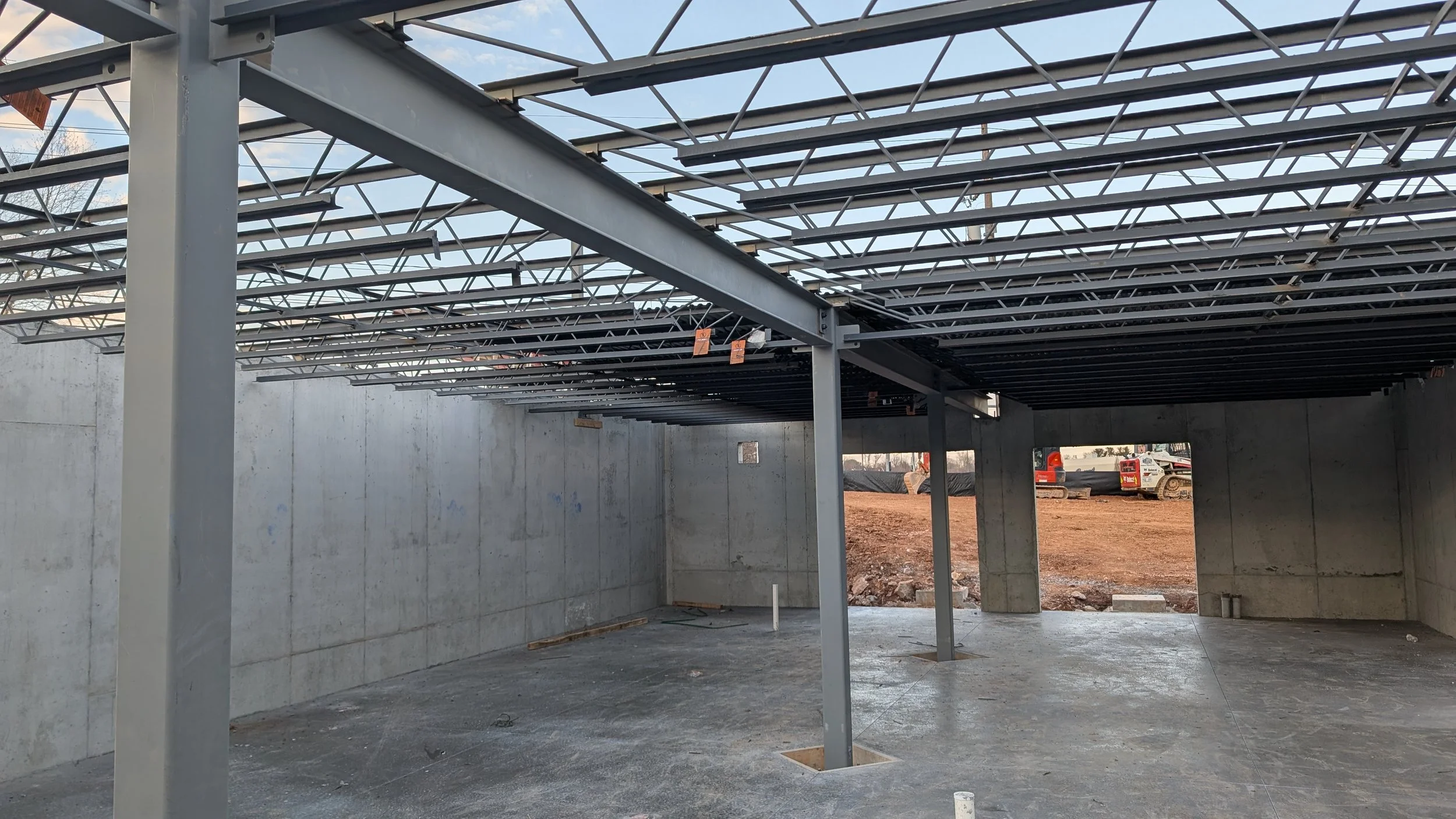 Construction site with steel framework and concrete walls, partially enclosed space, dirt outside, construction equipment visible in the background.