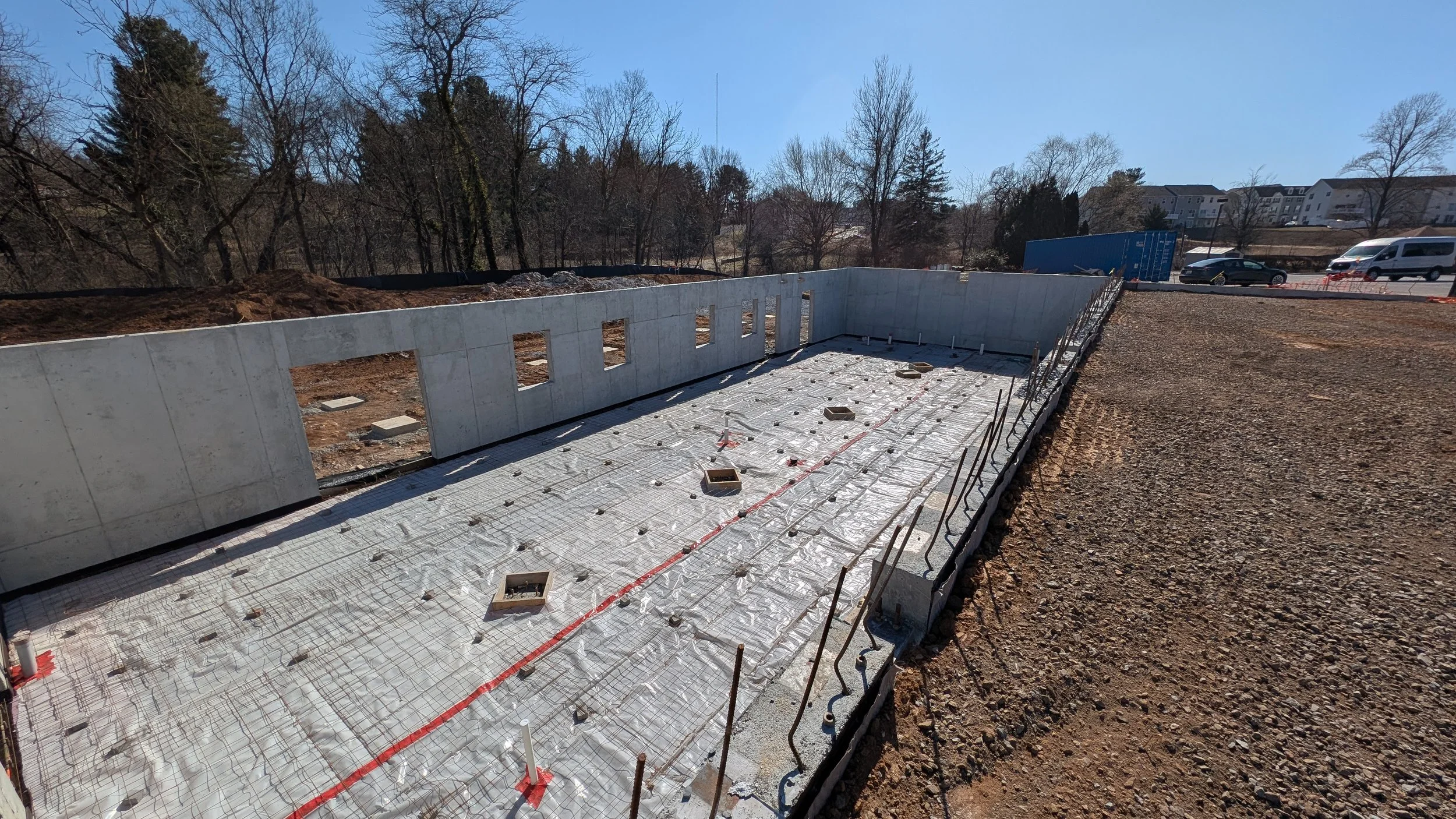 Construction site of a building foundation with concrete walls and rebar on the ground, surrounded by dirt and gravel, with trees and parked cars in the background.