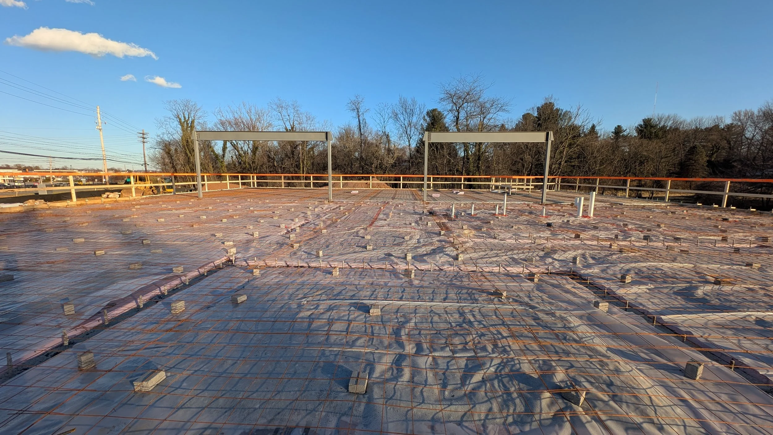 Construction site with a concrete slab foundation, metal rebar grid, and small vertical pipes, overlooking a tree line and blue sky.