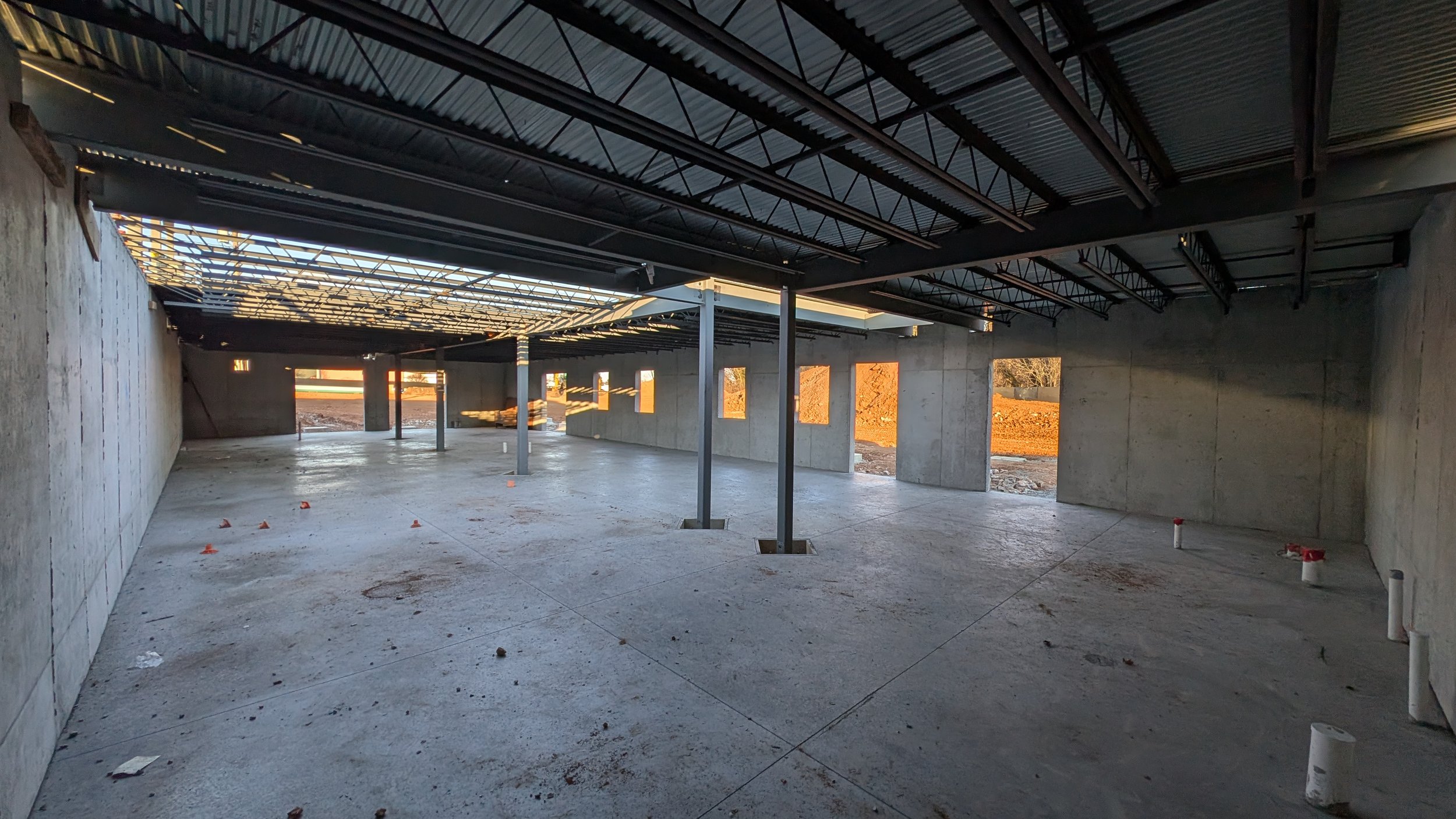 Interior view of a construction site with a concrete floor and unfinished walls, steel support beams, and an open ceiling with exposed metal roofing. Openings in the walls suggest windows or doors, with construction materials scattered on the ground.