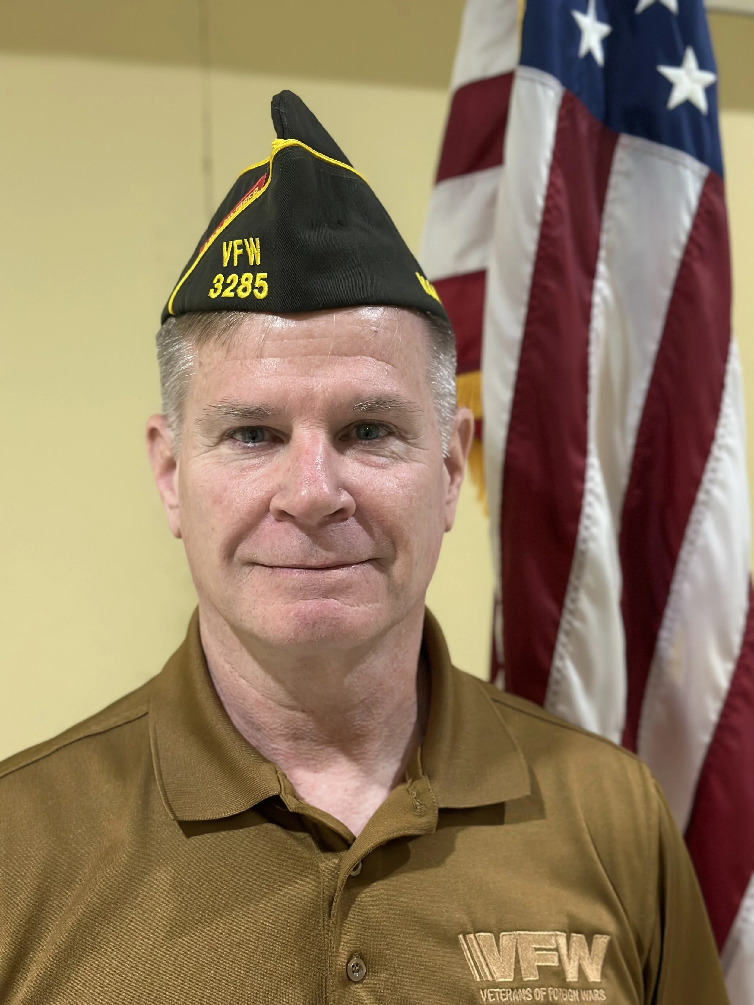 A man wearing a brown VFW uniform with a black cap labeled 'VFW 3285' stands in front of an American flag.