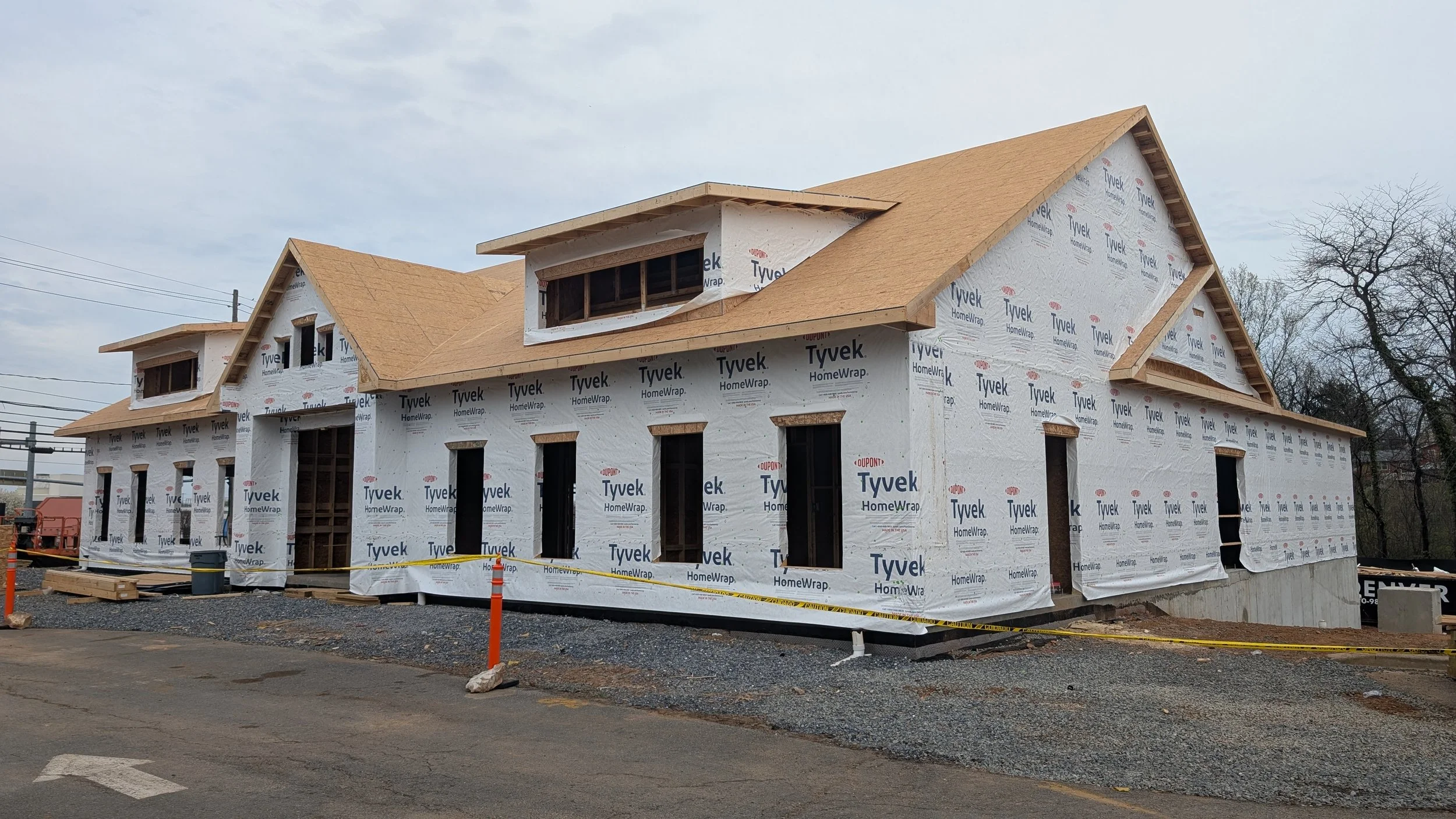A house under construction with wooden framing and a partially installed roof, wrapped in Tyvek HomeWrap, on a construction site with orange safety cones and gravel ground.