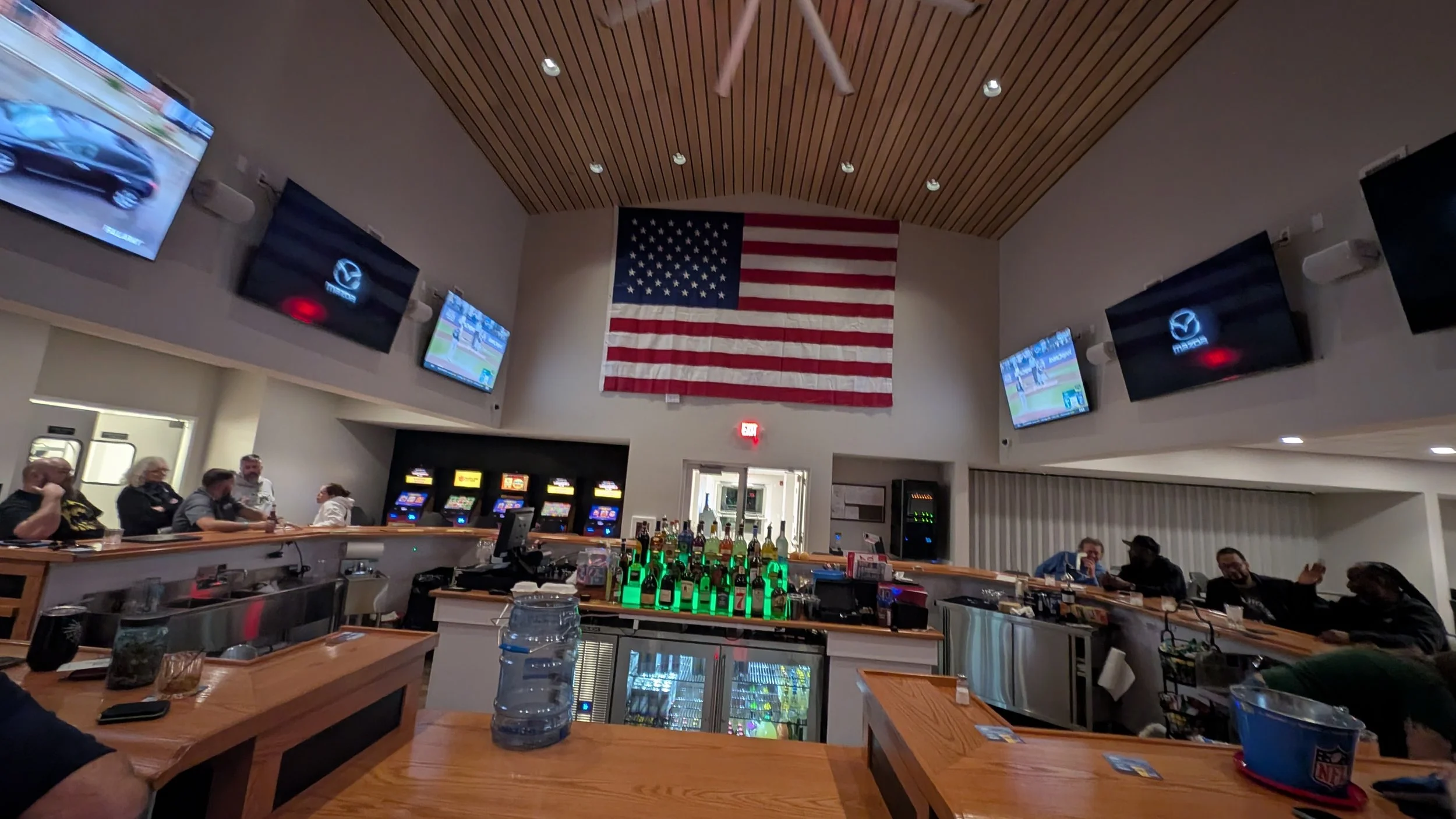 A bar area inside a restaurant with an American flag hanging on the wall, multiple televisions, and patrons seated along the bar and tables.