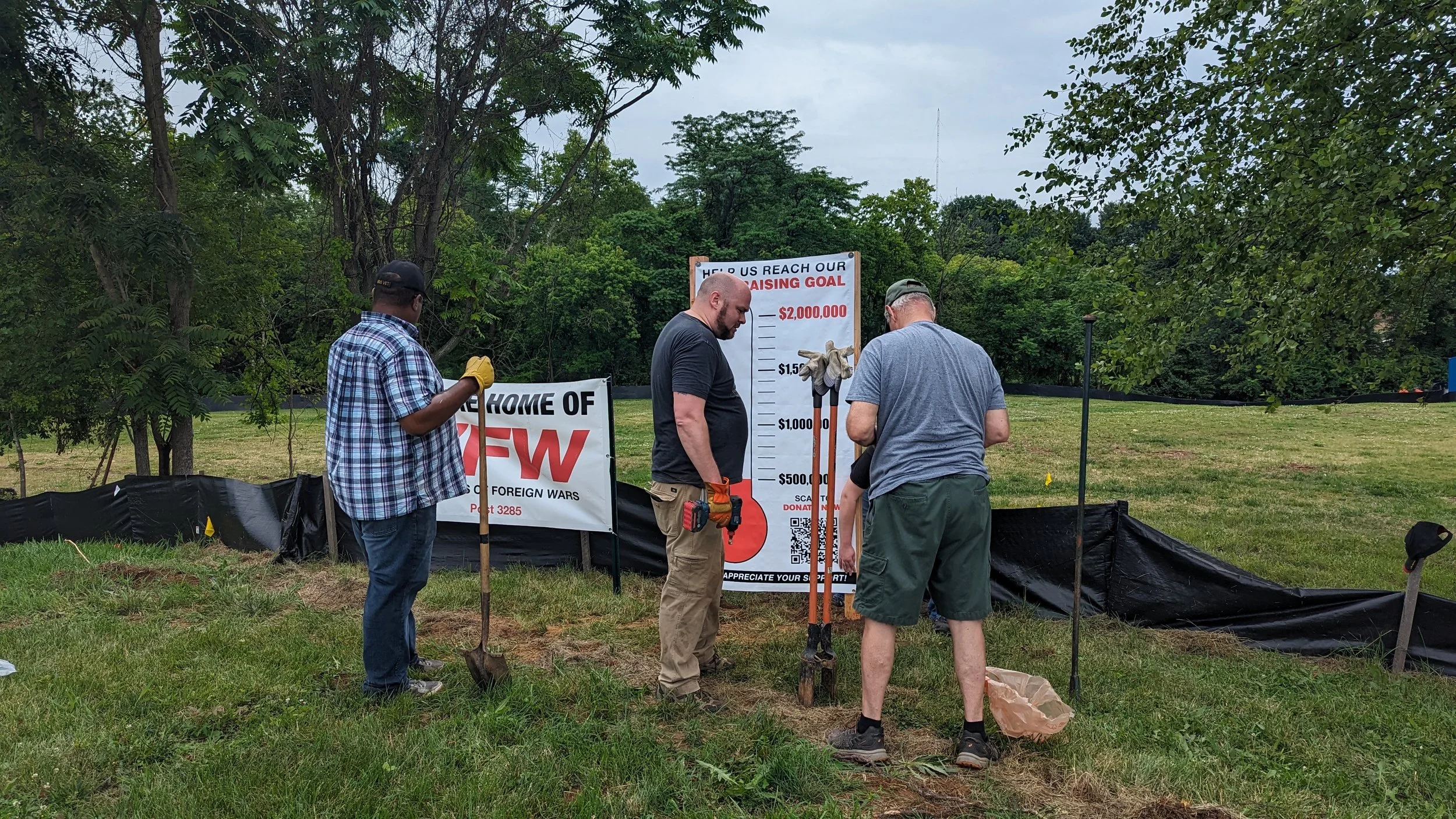 Four men planting a tree in a park with a sign asking for donations to reach a $2,000,000 goal, and a flag with the message 'Home of TFW'.
