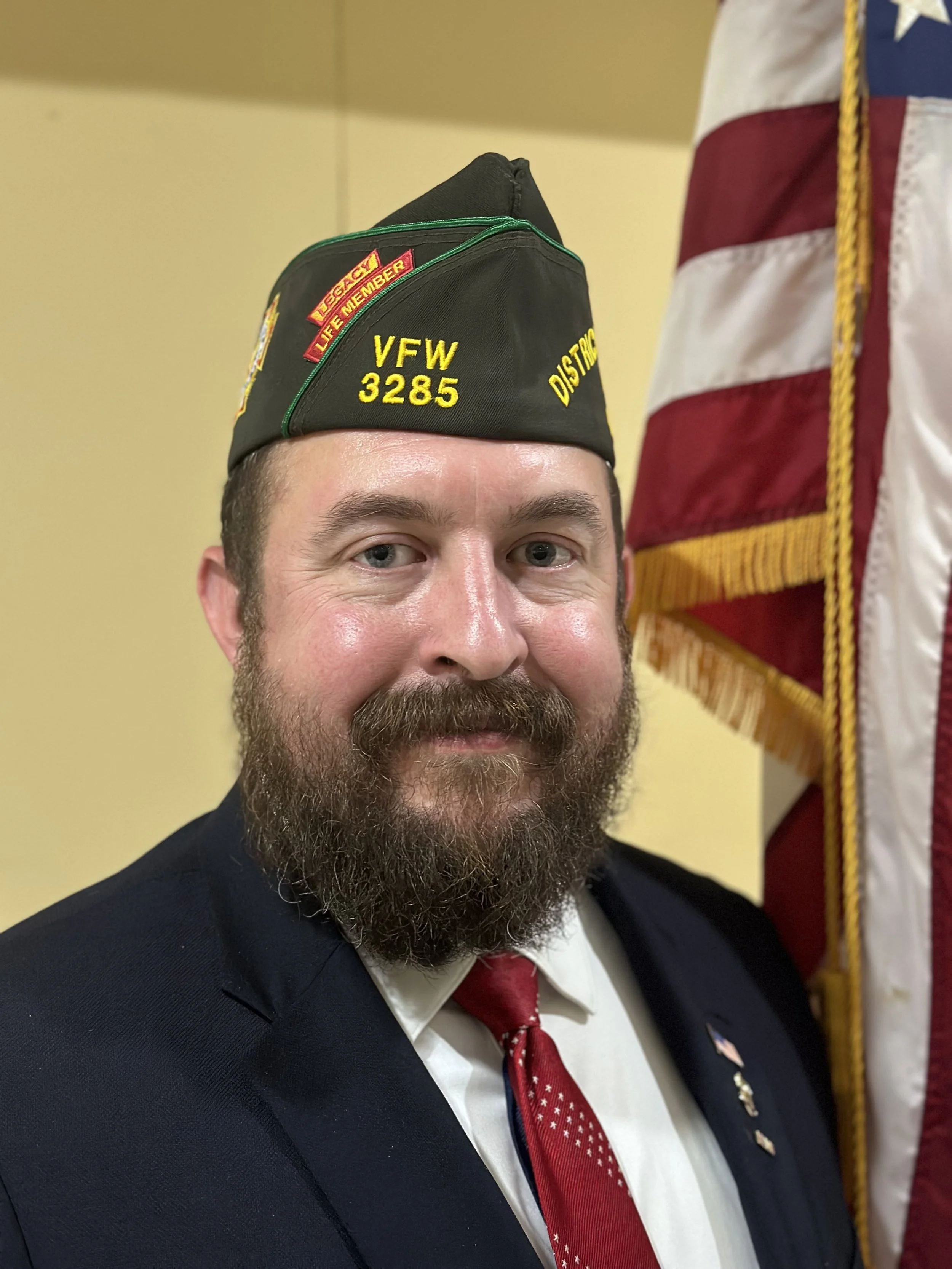 A man with a beard wearing a military-style hat, suit, and a red tie, standing next to an American flag.