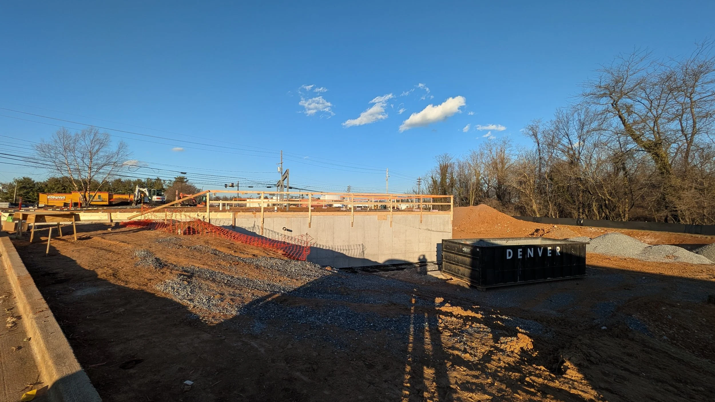 Construction site with a concrete wall and dirt piles, a black dumpster labeled 'DENVER', and several trees with bare branches in the background, under a blue sky with some clouds.