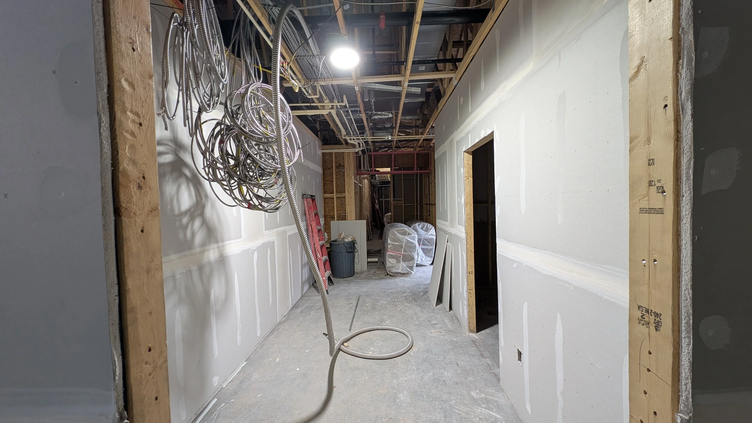 Interior view of a construction site with drywall installed on the walls, exposed wooden framing, hanging electrical wires, and building materials stored in the background.