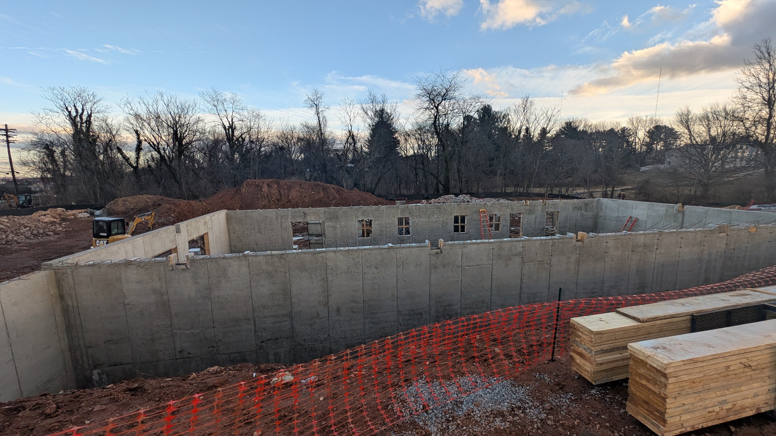 Construction site with concrete foundation walls, wooden stacks, and a small excavator, with a background of leafless trees under a blue sky with clouds.