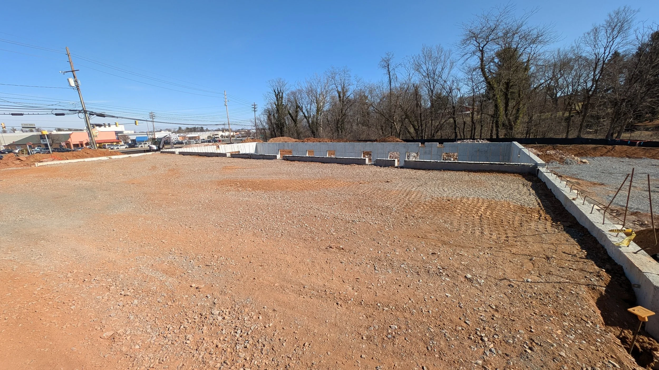 Construction site with a foundation in progress, gravel ground, and a concrete wall. Background includes trees, power lines, and buildings under a clear blue sky.
