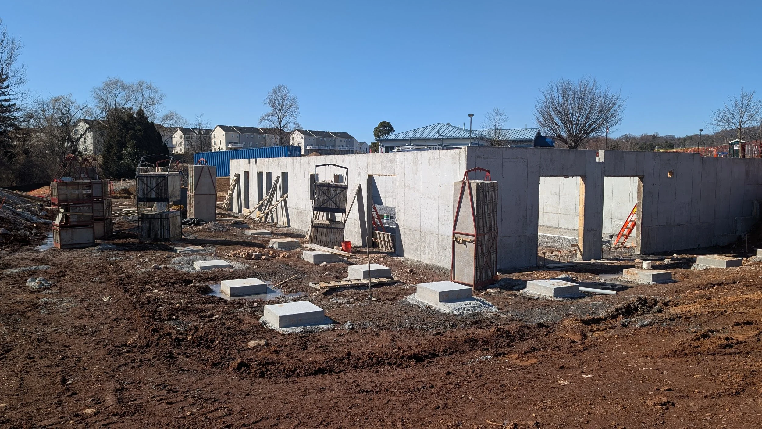 Construction site with concrete foundation wall and temporary structures for building support, surrounded by dirt and construction materials, under clear blue sky.