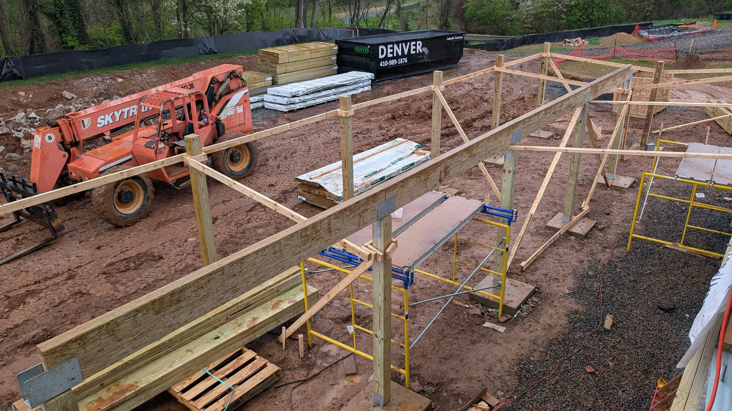 Construction site with wooden framework, scaffolding, dirt ground, and construction equipment including an orange telescopic loader and black containers.