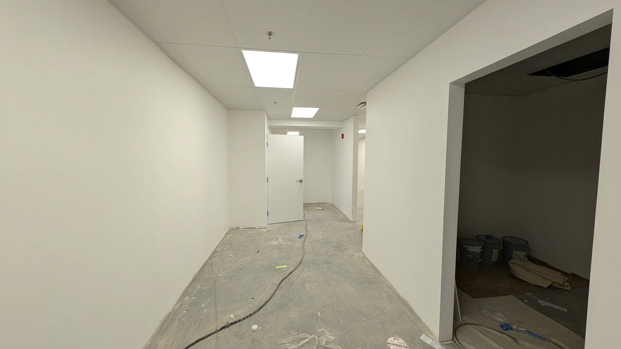 Interior of a building under construction with white walls, a suspended ceiling, and unfinished flooring. There are construction materials and tools visible, and some rooms or spaces are partially open.