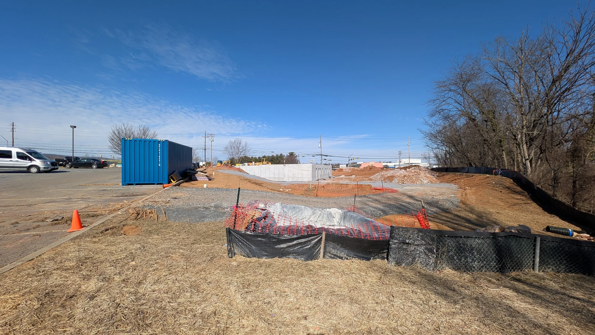 Construction site with dirt piles, orange safety fencing, a blue storage container, and parked cars in the background on a clear day.