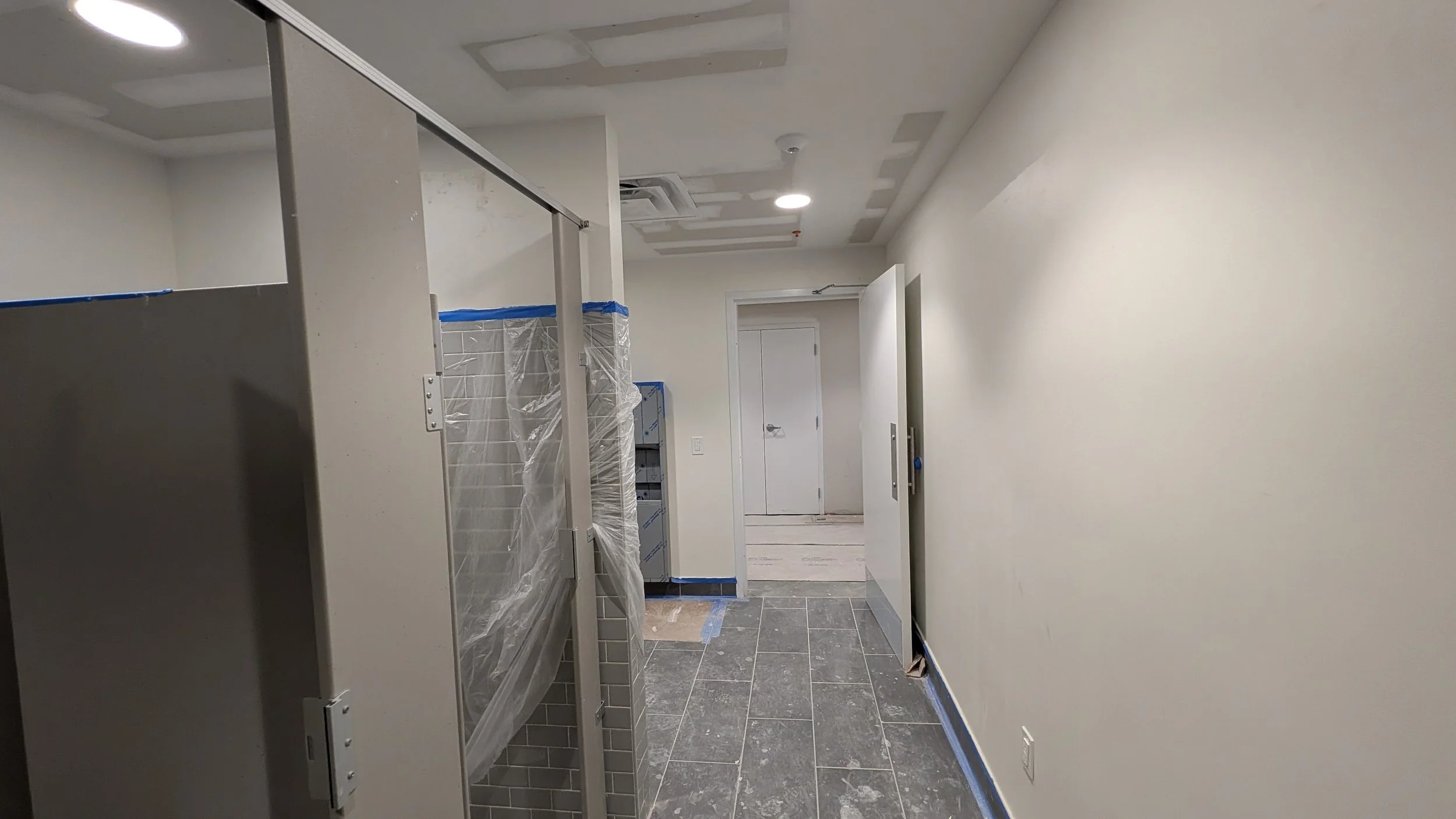 Interior view of a corridor under construction or renovation, with unfinished ceiling and walls, portable lockers covered in plastic, and a doorway at the end.