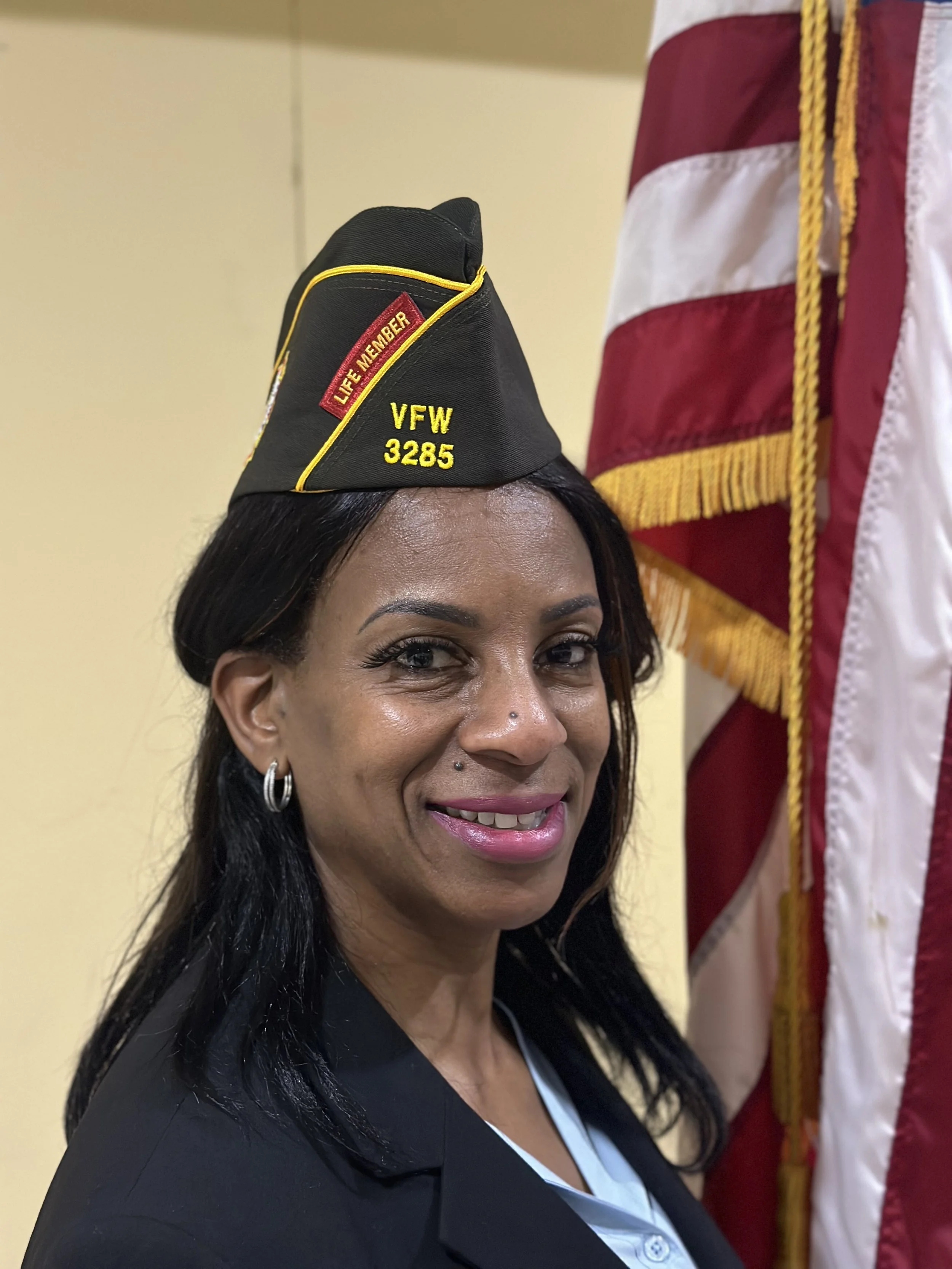 A woman with black hair wearing a VFW cap, black blazer, and earrings, standing next to an American flag, smiling at the camera.