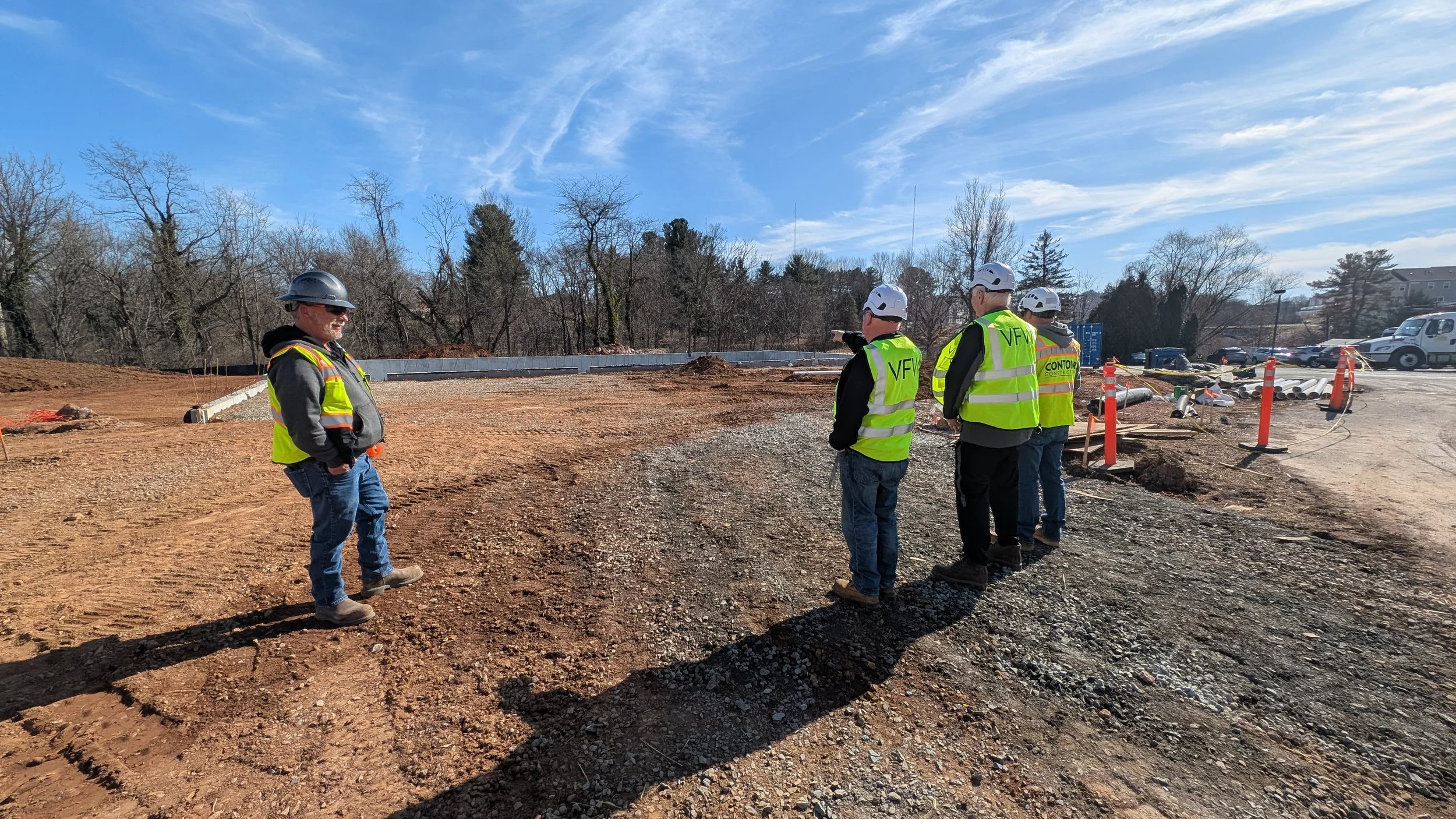 Group of construction workers in safety vests and helmets inspecting a construction site with dirt ground, pipes, and construction cones under a blue sky with scattered clouds.