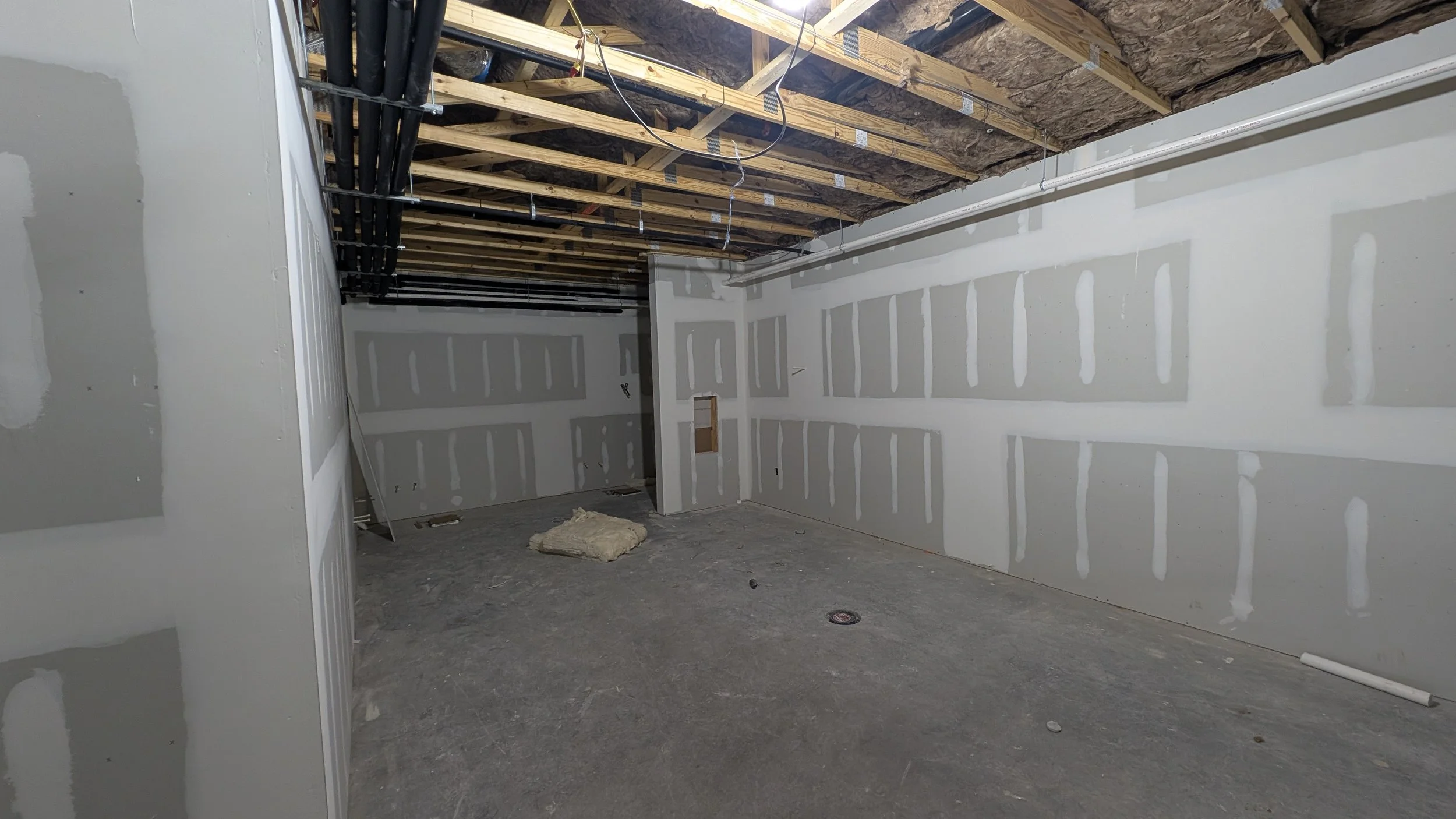Room under construction with drywall installed on the walls, exposed ceiling framing and electrical wiring, and construction materials on the floor.