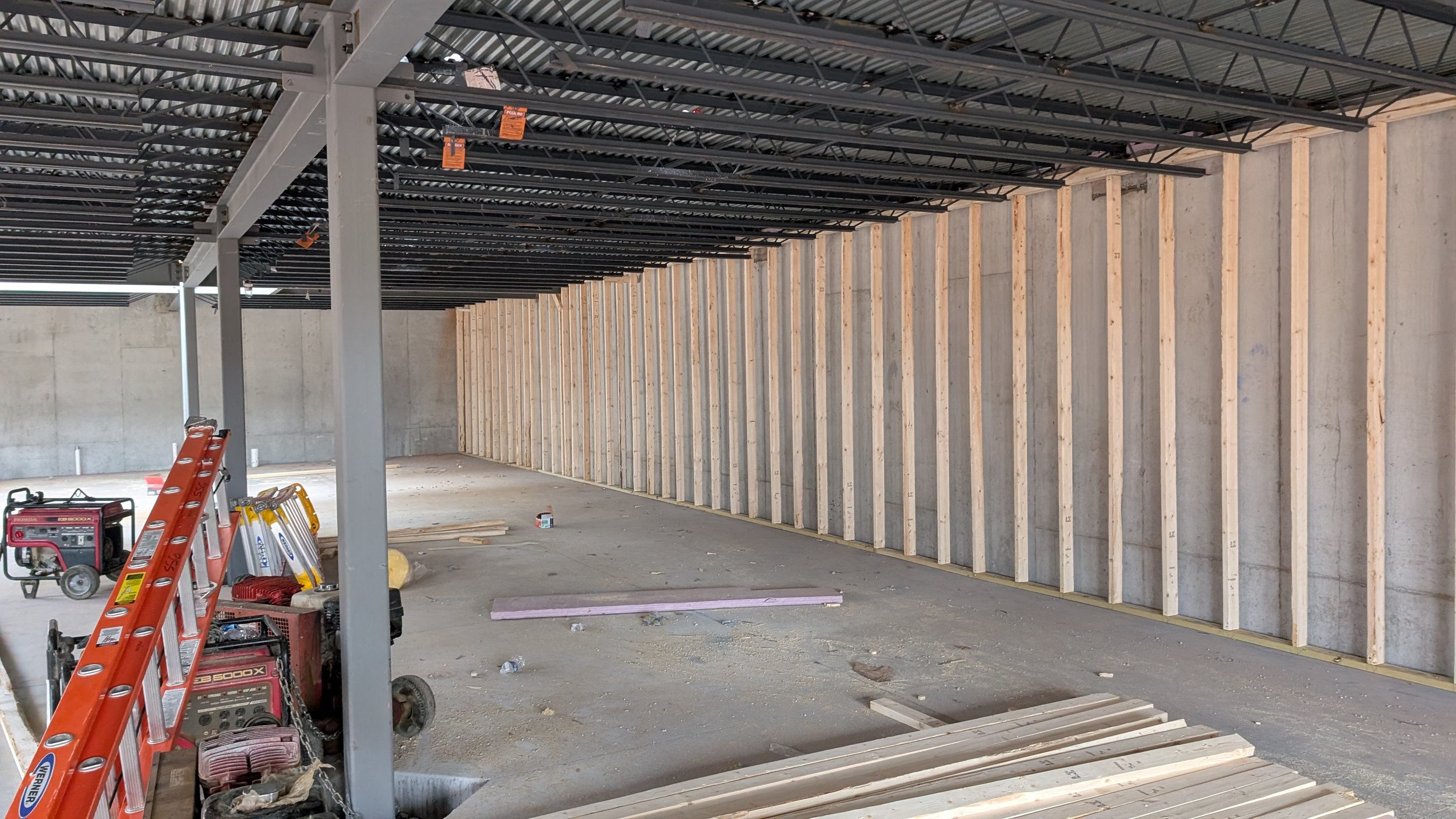 Interior view of a building under construction with wooden framing along the walls, unfinished concrete floor, construction tools, and materials scattered around.