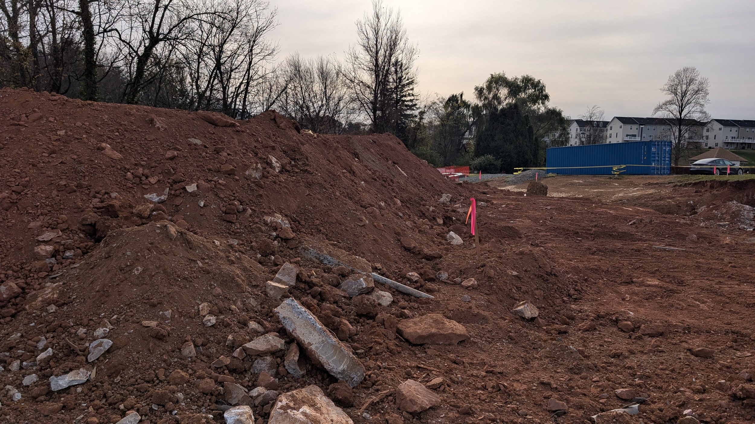 Construction site with large dirt piles, scattered rocks, pink safety markers, a blue shipping container, and distant residential buildings under an overcast sky.